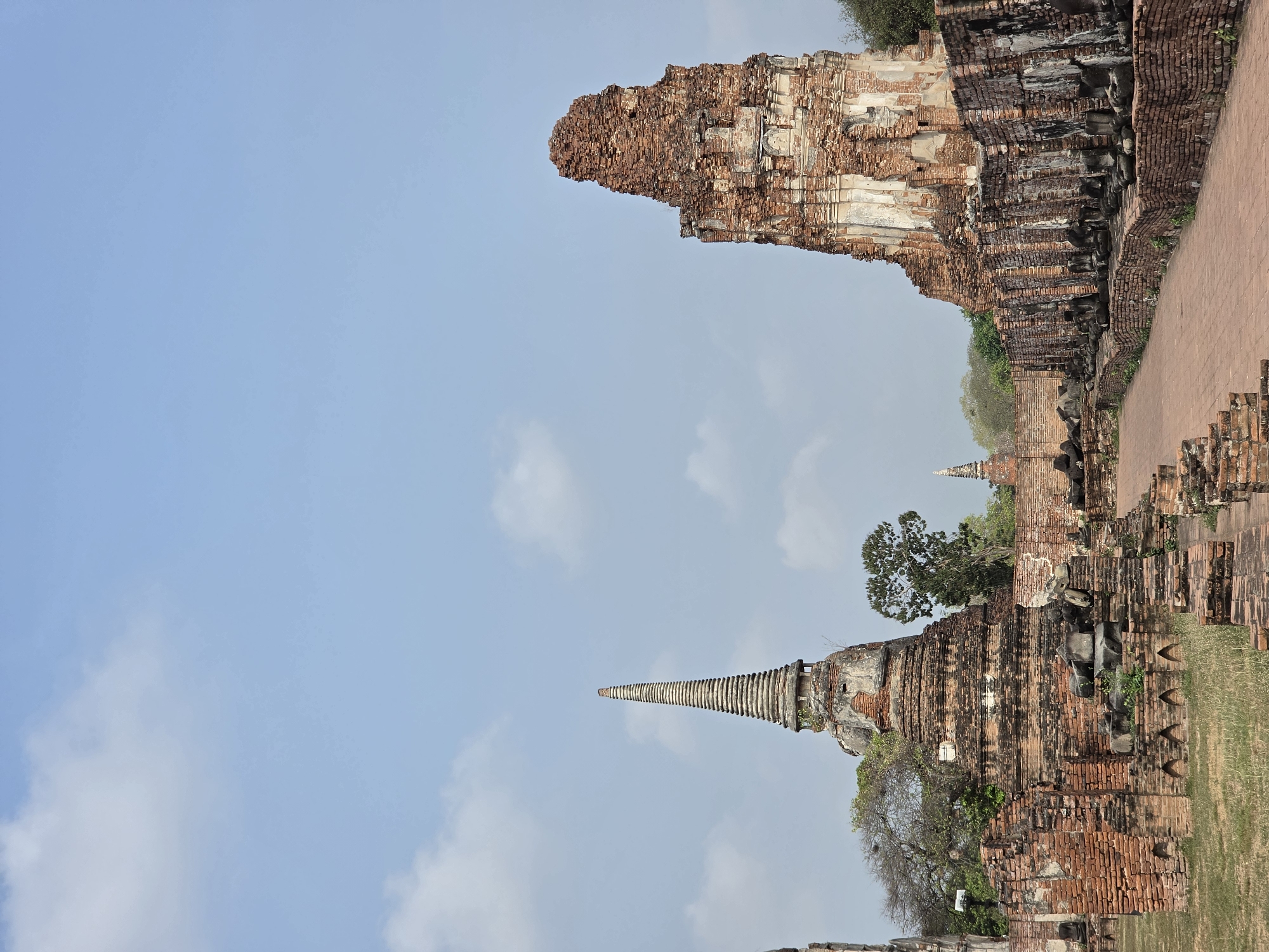 The image captures a landscape with historical ruins in Phra Nakhon Si Ayutthaya City Municipality, Thailand. The ruins, primarily constructed of red brick, are scattered throughout the landscape. There are no people present. The sky is partially cloudy, and the weather appears clear. Some of the structures consist of crumbling walls and towers, while others are more intact. The environment suggests a site of historical or cultural significance, now open to the elements.