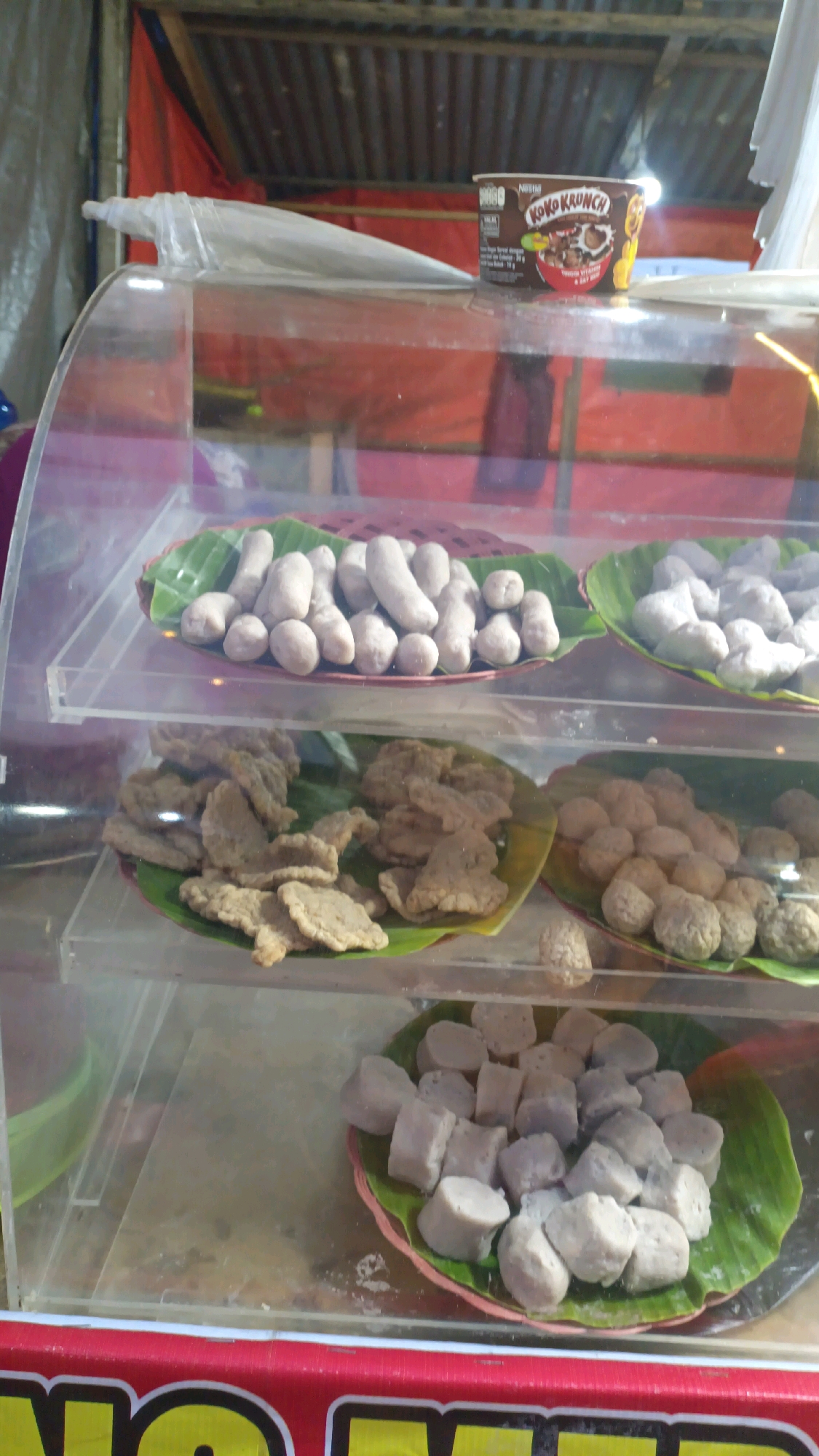 The image shows a food display case at a street food stall. In the upper section of the display, two plates of round, light-colored food items, likely meatballs or similar snacks, are visible. These are placed on banana leaves. Above the display case, a cup of "Koko Krunch" cereal is also present. The middle section of the display contains two plates of flat, irregular-shaped fried food items, also on banana leaves. The bottom section of the display features a plate of cubed food items, again on a banana leaf, which appear to be some type of fish cake or similar preparation. The food stall appears to be outdoors, with a corrugated metal roof and red tarpaulin visible in the background, suggesting a bustling market or roadside setting in Padang Panjang, Indonesia. The overall impression is of a local eatery offering a variety of prepared foods.