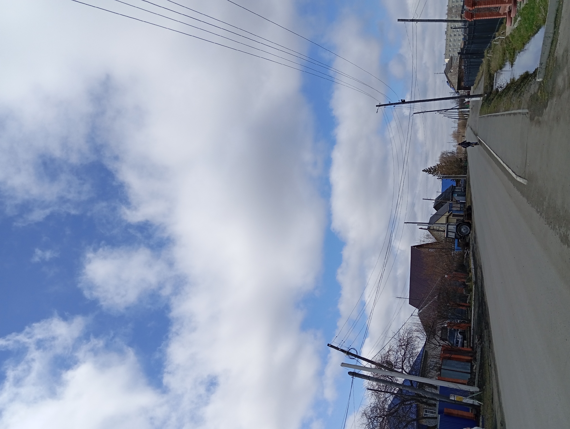 The image shows a street in Yurgamysh, Russia. The street runs vertically through the frame, with buildings lining the right side and a mix of sky and clouds filling the left. A road with a cyclist is visible along the side. Power lines stretch across the upper portion of the image, contrasting against the sky. The weather appears overcast with a mix of blue sky visible among the clouds. Buildings are visible in the background.