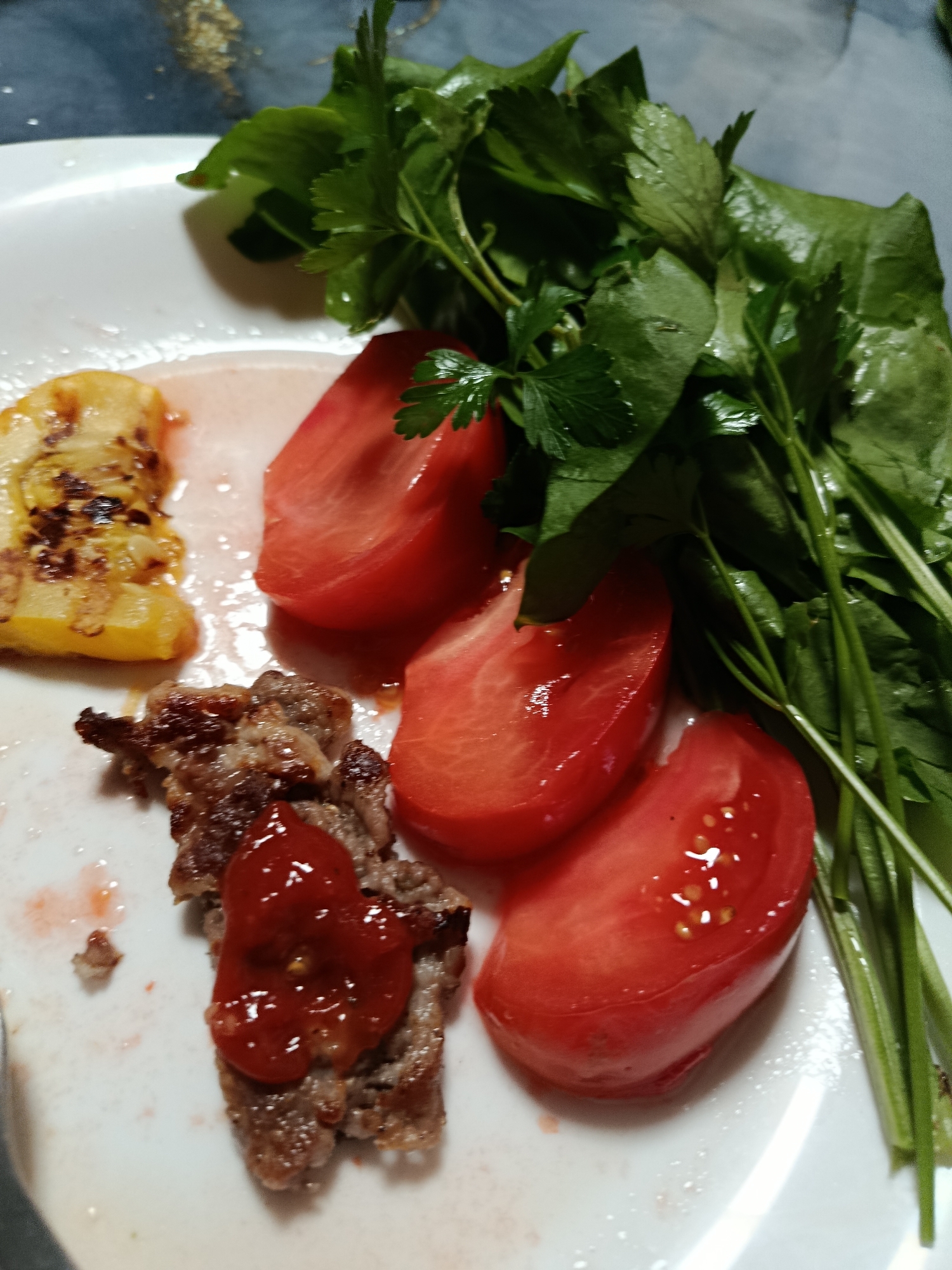 A plate of food is presented on a white surface. The plate contains a piece of grilled meat topped with ketchup, slices of red tomato, a piece of grilled yellow bell pepper, and a bunch of fresh green herbs. The food is arranged on the plate in a way that suggests a meal is about to be consumed.