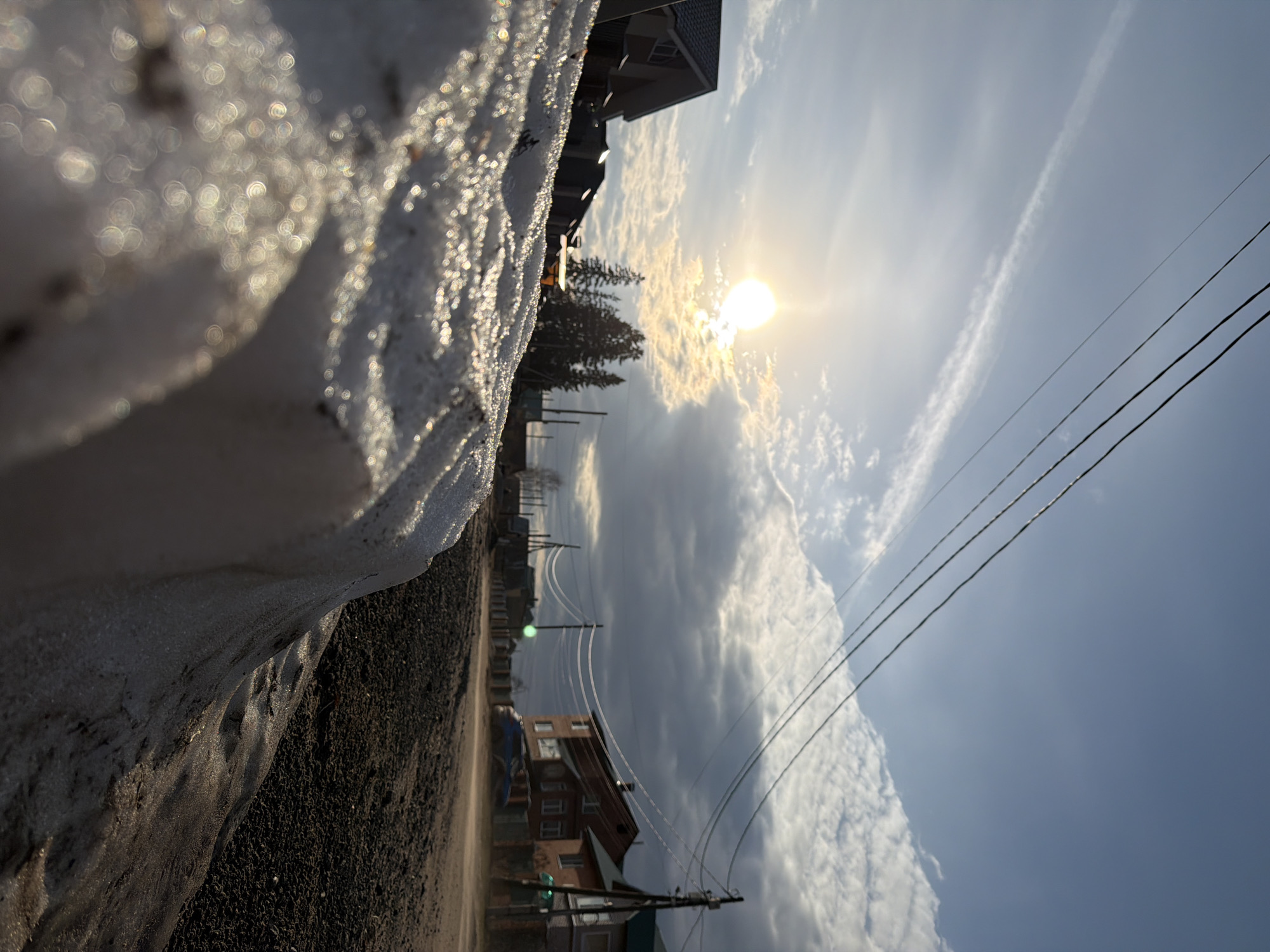 This media file presents a street-level view on a bright, cold day, likely in Chaikovsky, Russia. The image is taken from a very low perspective, looking forward and slightly upwards, suggesting it was captured near the ground.
In the immediate foreground, a bank of sparkling snow or ice runs along the bottom left edge of the frame. The surface of the snow glistens with numerous bokeh-like reflections from the sunlight, indicating a crystalline or icy texture. This icy edge borders what appears to be an unpaved dirt or gravel road that stretches into the midground.
Along the road, several utility poles with multiple overhead wires crisscross the scene, receding into the distance. On the left side of the road, evergreen trees are visible, behind which stand residential buildings with multiple stories and visible windows. More houses are scattered further down the road, contributing to the sense of a populated, possibly rural or suburban, area.
The background is dominated by the sky, which transitions from a bright, slightly hazy blue to areas with patchy white and grey clouds. The sun is a powerful, bright orb in the upper right quadrant of the sky, partially obscured by clouds but casting a strong light across the scene. Several thin, straight contrails are etched across the upper sky.
The time of day appears to be either morning or late afternoon, given the strong, somewhat low angle of the sunlight. The clear sky with clouds and the prominent snow/ice indicate cold weather, characteristic of winter or early spring. No people or specific activities are depicted, and there is no visible text in the image.