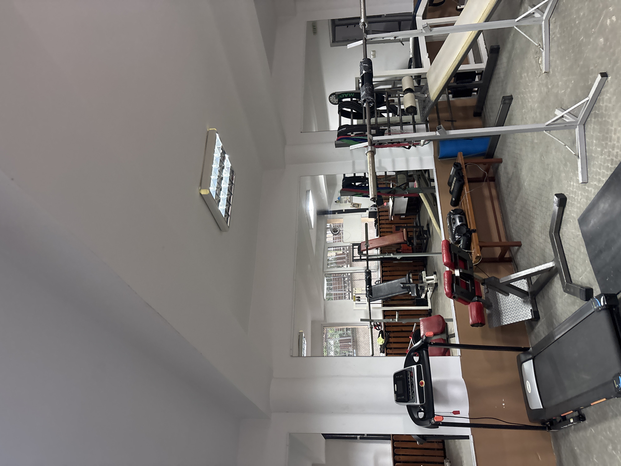 The image shows the interior of a gym, likely in Zlatograd, Bulgaria, given the prompt. The gym features a variety of exercise equipment, including a treadmill in the foreground, various weight machines, and a squat rack in the background. The gym appears to be well-lit with white walls and a mirrored wall to create the illusion of more space. There are no people visible in the frame.