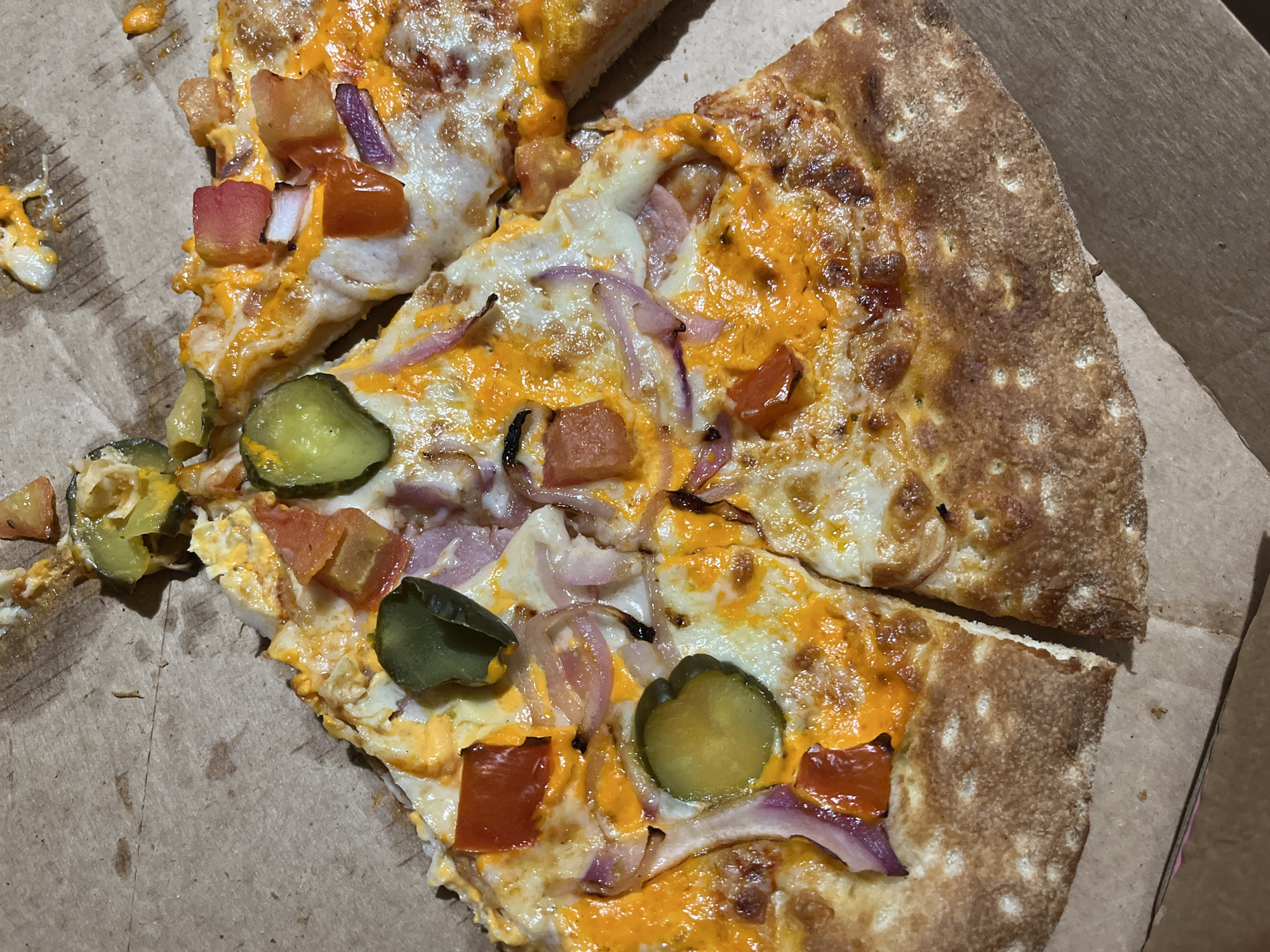 The image shows slices of pizza on a cardboard pizza box. The pizza appears to be topped with cheese, tomatoes, red onion, and pickles. The pizza is golden brown and appears to be cooked. The pizza box is a light brown color and shows some signs of wear. The location is not specified in the image.