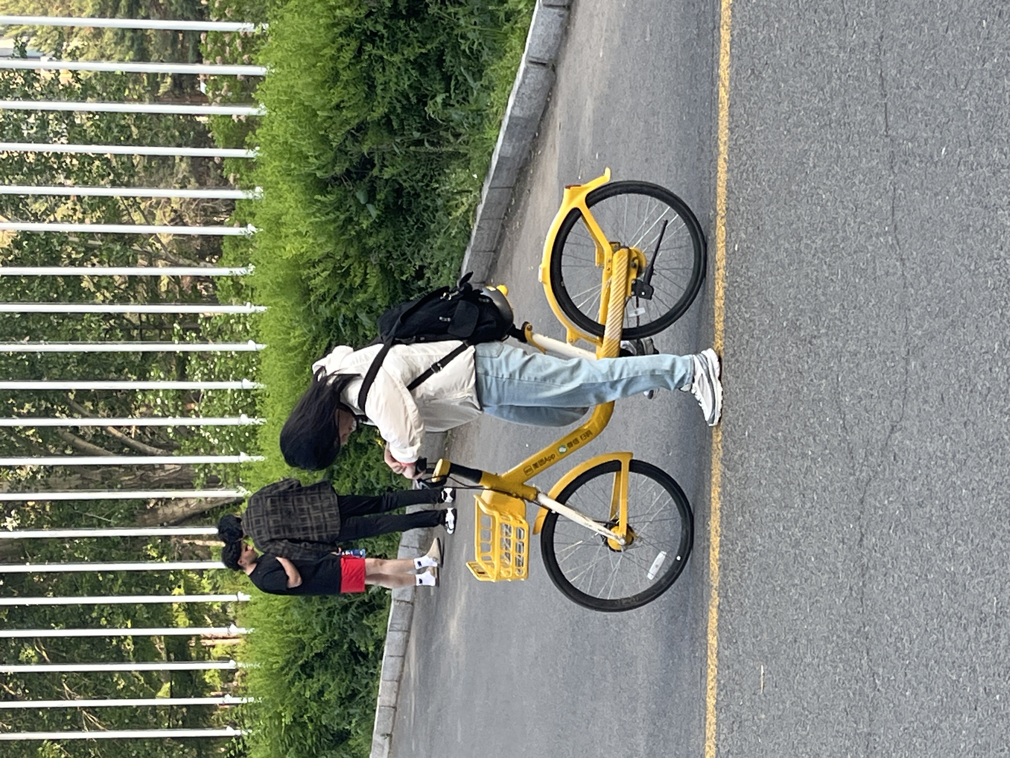 A yellow bicycle is parked on an asphalt road alongside a curb in Changchun City, China. A young woman with long black hair, wearing a white jacket and light blue jeans, stands beside the bicycle. She is carrying a black backpack. Nearby, two figures are partially visible, walking on the pavement adjacent to a hedge and a white fence. The road has a yellow line marking its edge. The environment suggests a park or campus setting with greenery and pedestrian walkways.