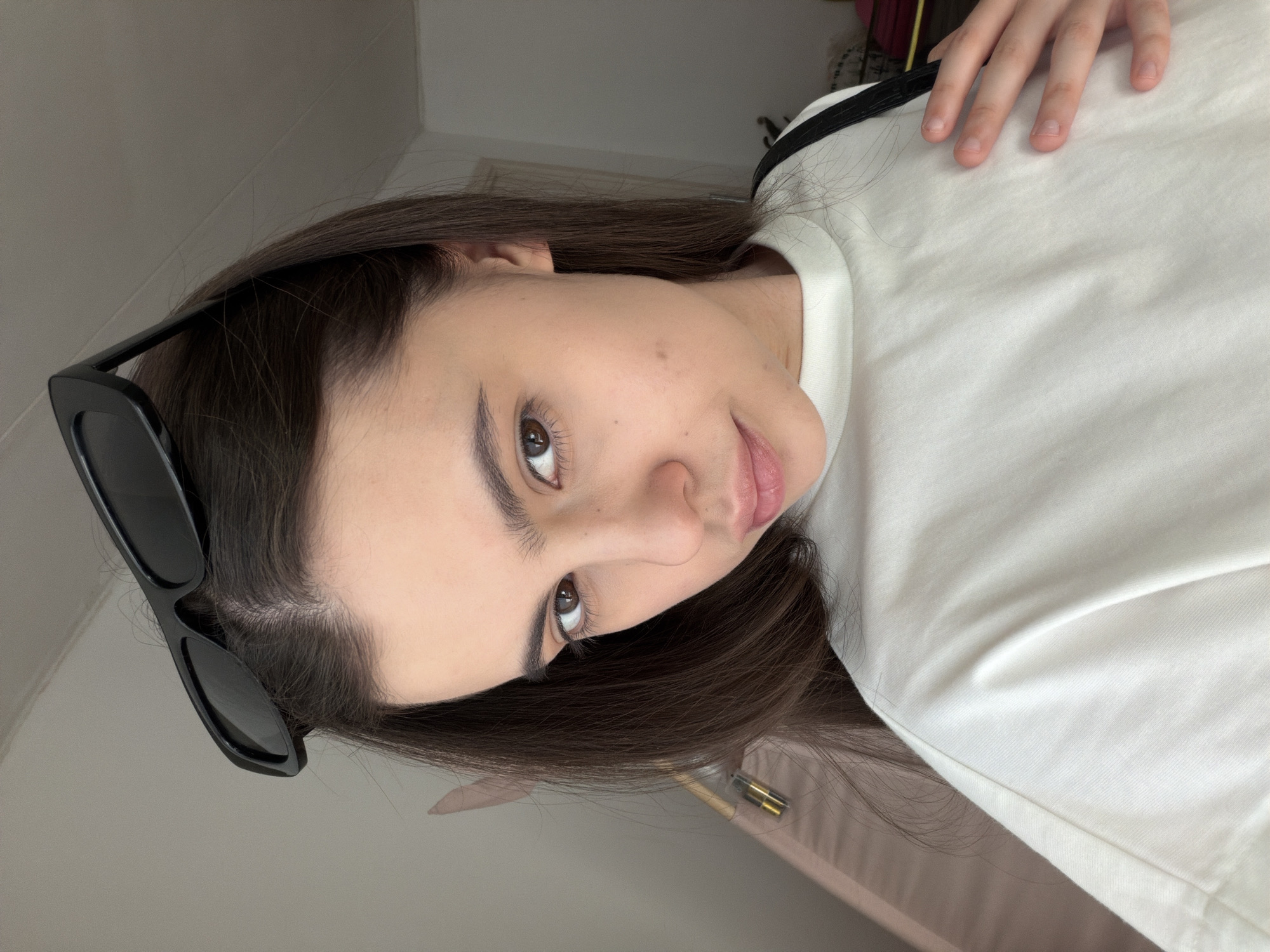 A woman is posing for a close-up selfie indoors. She is wearing a white t-shirt and has sunglasses perched on her head. She has brown hair and brown eyes. The location cannot be determined from the image, and the city of Chaikovsky, Russia, is not identifiable.