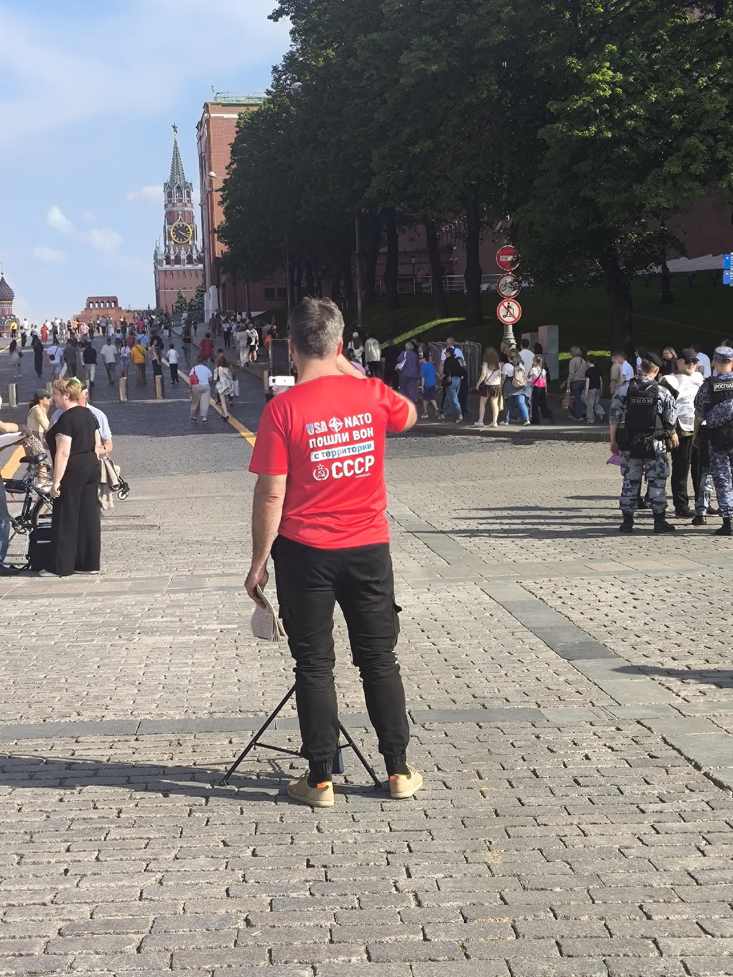 A man in a red T-shirt with Russian writing stands on a cobblestone square, likely making a video with a smartphone on a tripod. Several people are walking in the background. Also, there are several officers, likely police, standing on the edge of the square. In the background, the Spasskaya Tower, a landmark of Moscow, Russia, is visible. The sign on the back of the shirt reads, in translation, “USA, NATO go away from the territory of the USSR.”