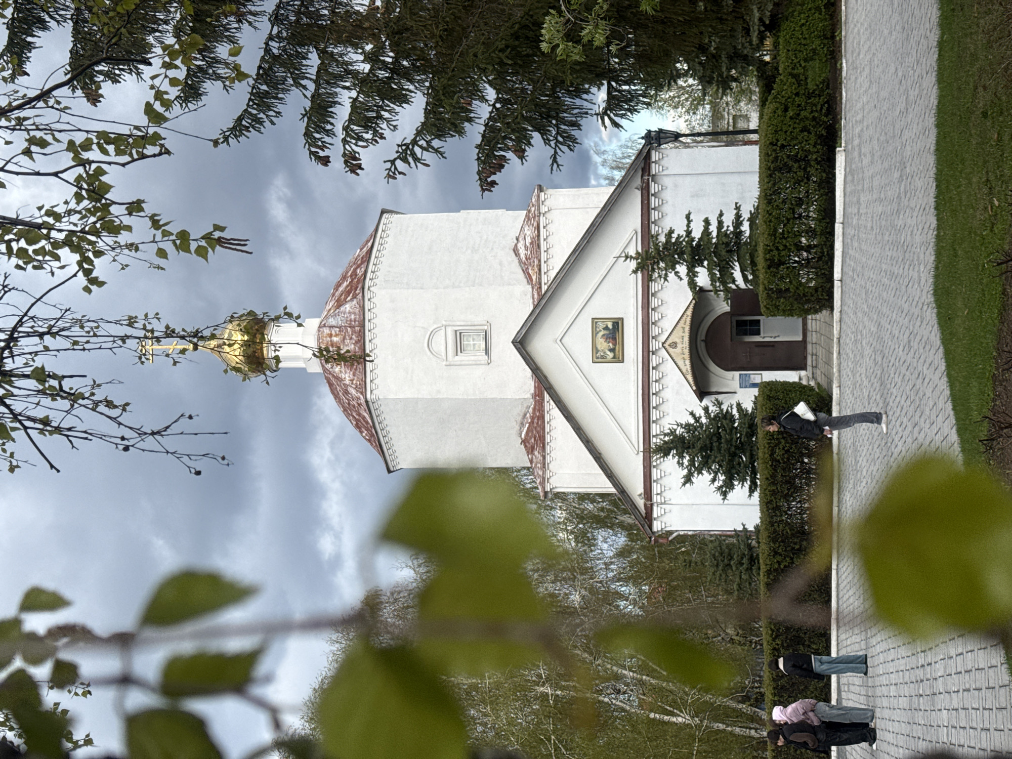 The image shows a white church in Syzran, Russia, viewed through tree branches. The church has a gold dome topped with a cross. The sky is cloudy. People are walking along a paved path adjacent to the church. A trimmed green hedge separates the path from the church grounds.