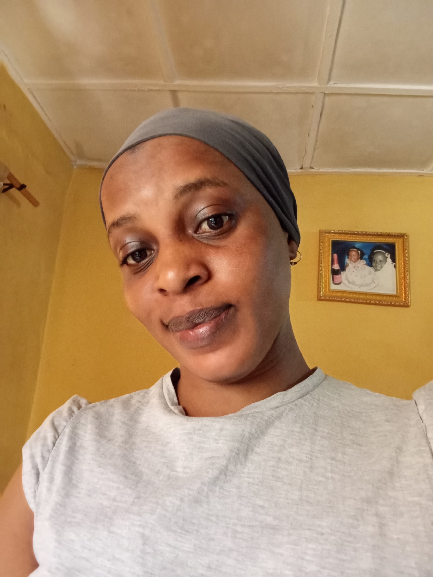 A medium close-up shot shows a woman with dark brown skin wearing a gray headwrap and a gray T-shirt. The background is a yellow wall with a framed picture hanging on it. The room is in Narayi, Nigeria. The picture in the background features two young children with a cake and a bottle of wine.