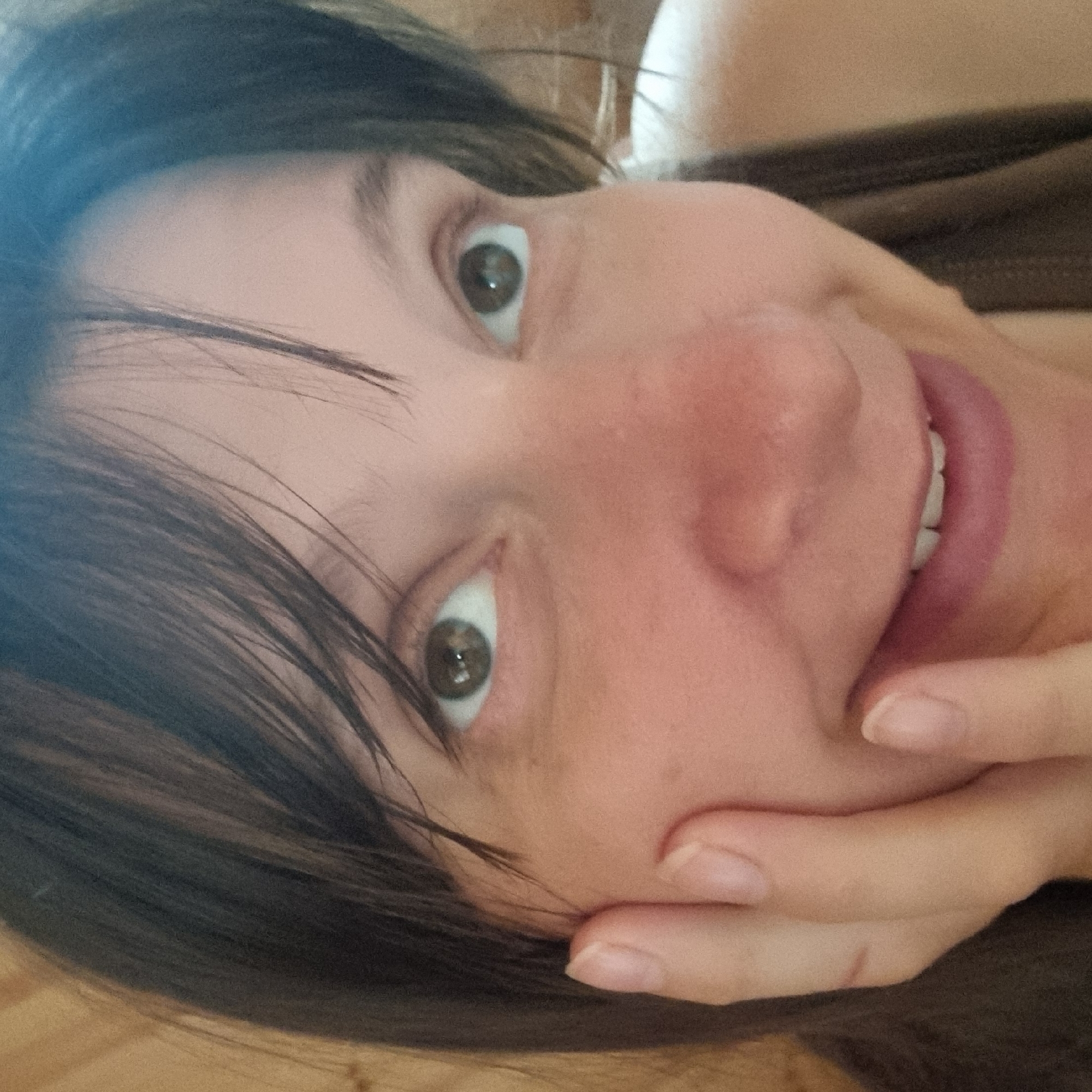 A close-up, eye-level, medium shot shows a woman's face, from the nose up, with her left hand resting on her cheek. She has brown hair that partially obscures her forehead and is looking slightly to her left, with her mouth open in a smile. Her eyes are green and her skin is fair. The background is blurred, with hints of wood grain, suggesting an indoor environment in Dunnae-myeon, South Korea.