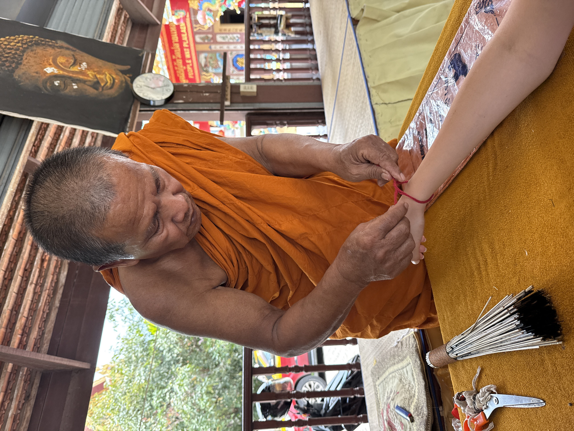 A Buddhist monk, dressed in an orange robe, is in the process of tying a red string bracelet onto the wrist of a person's arm. The monk's hands are carefully manipulating the string. The person's arm, which appears to be that of a child or young adult, is resting on a yellow cloth-covered surface.
In the foreground, to the left of the person's arm, are a pair of orange-handled scissors tied with string and a long object resembling a brush or whisk made of thin metal rods with black bristles at the end. A blue lighter is also visible on a woven mat.
The background shows a covered walkway with a wooden roof and railings. A painting of a Buddha head is visible on the wall. A clock is hanging on the wall below the painting. Further back, some red and gold structures with intricate designs are visible, suggesting a temple or shrine setting. Trees and foliage are visible outside the walkway, and a red vehicle, possibly a taxi, is partially visible through the railings.
The scene takes place outdoors under a covered structure, likely within a temple complex in the Ko Samui District of Thailand. The overall atmosphere appears to be one of ritual or blessing.