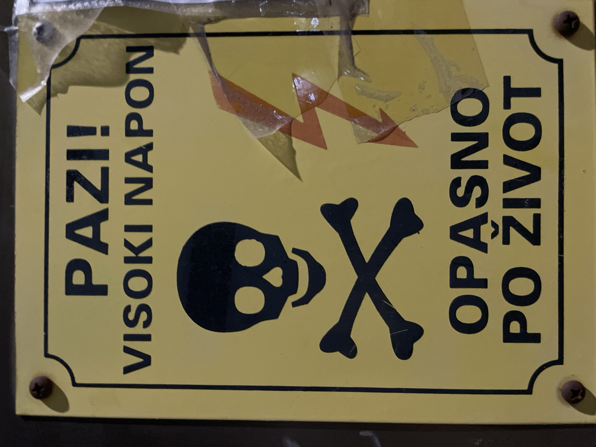 A rectangular, yellow warning sign with a black border and rounded corners is depicted, affixed to a dark, indistinct background by at least two visible dark screws, one in the top-left corner and another in the bottom-right. The sign features black capitalized text and symbols. At the top, the text reads "PAZI!" followed by "VISOKI NAPON". Below this, a black skull and crossbones symbol is prominently displayed on the left side of the sign. To the right of the skull and crossbones, an orange and red stylized lightning bolt symbol is partially obscured by torn, semi-transparent adhesive tape that covers the right edge of the sign. At the bottom of the sign, the text reads "OPASNO PO ŽIVOT". The sign appears worn, with minor surface abrasions and the aforementioned tape. No people or specific actions are visible. The environment is an unknown location within the city of Unknown, Unknown.