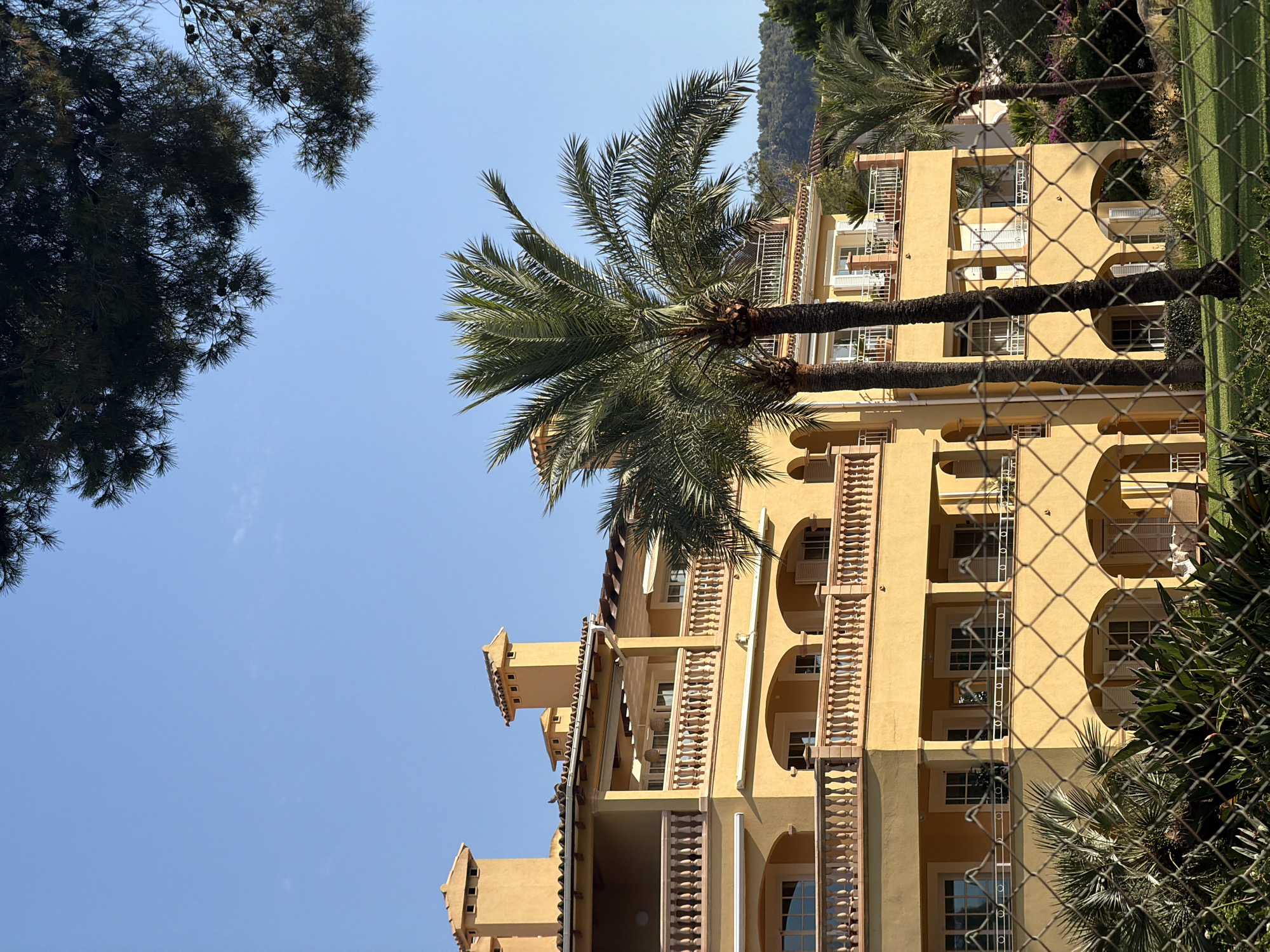 A multi-story, yellow-ochre building with balconies and terracotta tiled roofs stands under a clear blue sky. Two large palm trees are positioned in front of the building, with a wire fence in the foreground obscuring some of the lower portion of the structure and surrounding garden. The building appears to be a residential or hotel complex located in Calvià, Spain, suggested by the Mediterranean architectural style and palm trees. The scene is bathed in natural light, indicating a daytime setting, likely outdoors.