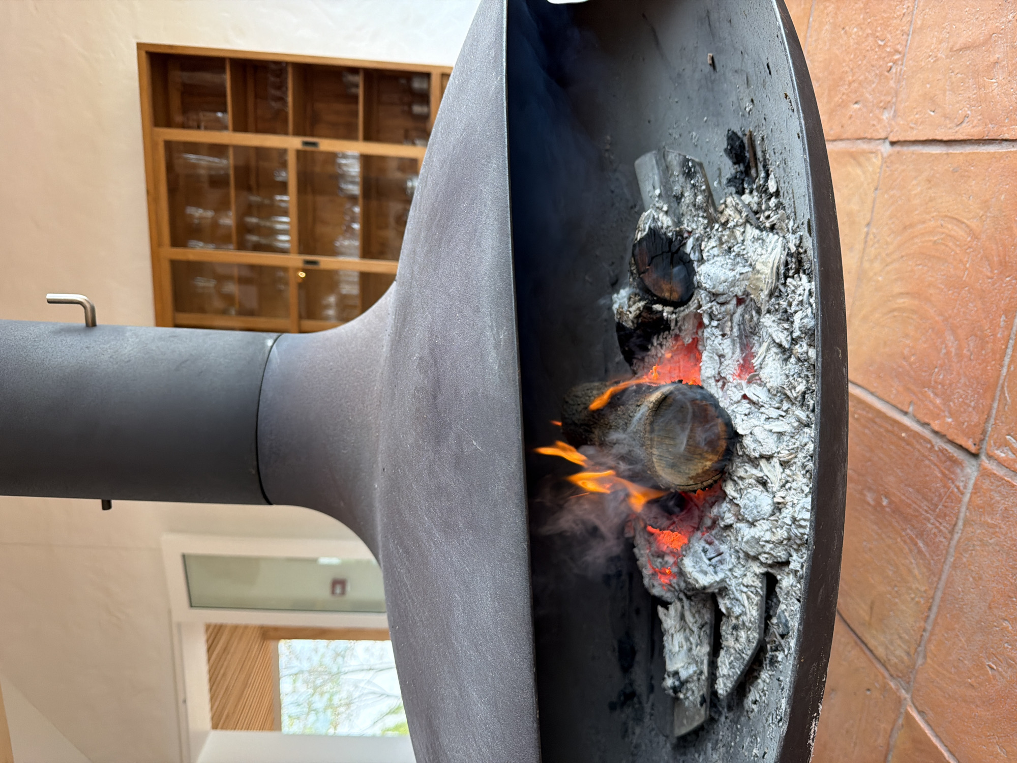 The image shows a modern, round, black metal outdoor fireplace in Hout Bay, South Africa. The fire is lit with wood burning inside, covered with ash and glowing embers. The fireplace is situated indoors near a wall tiled with orange bricks. A modern window and a wooden cabinet with glass doors can be seen in the background.