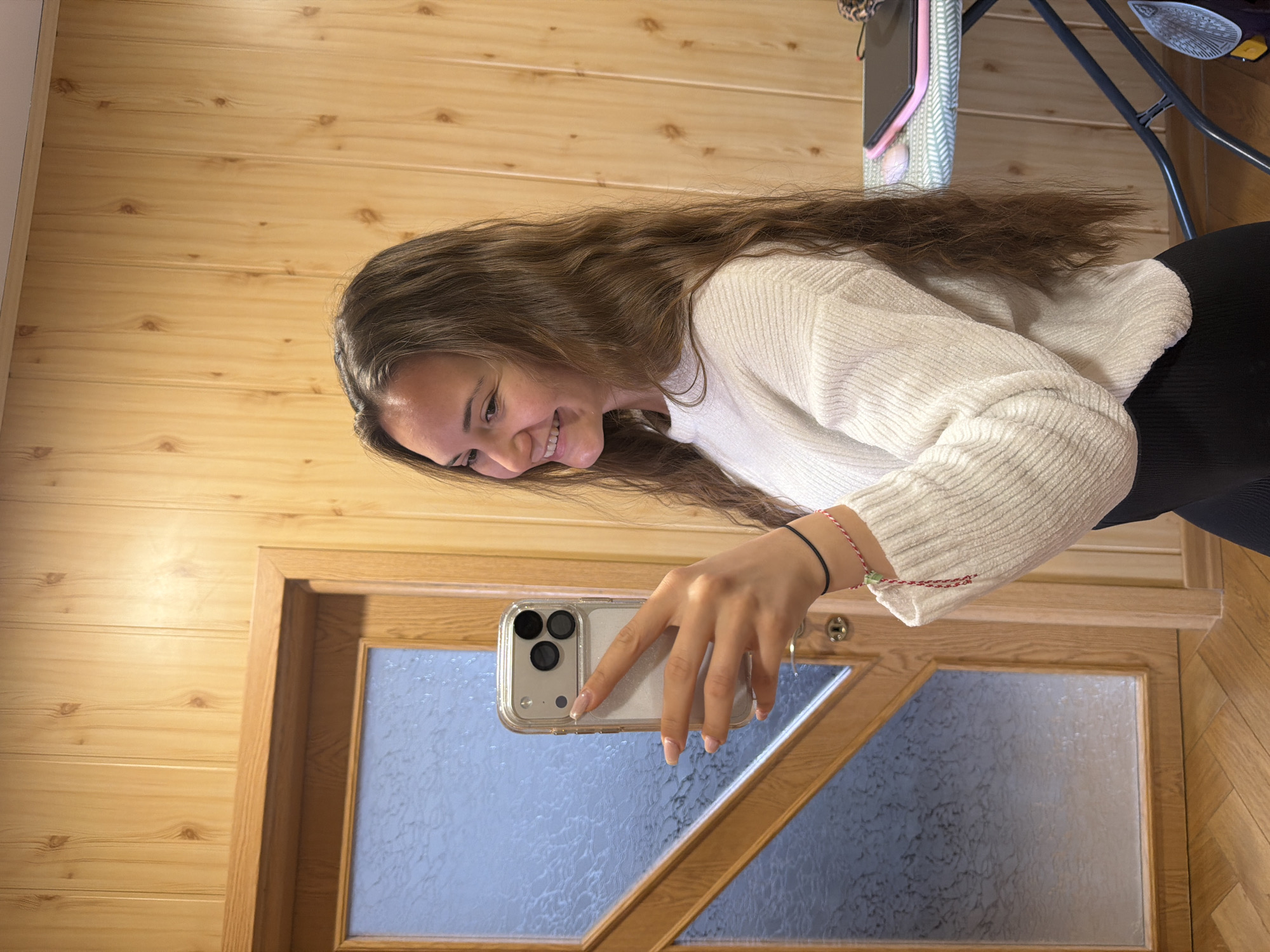 A young woman with long, wavy brown hair is taking a selfie with a smartphone. She is smiling and wearing a white sweater and black bottoms. She stands in front of a wooden door with a frosted glass panel, inside of a room with wood paneled walls. A table with items sits in the background. The location is Buhovo, Bulgaria.