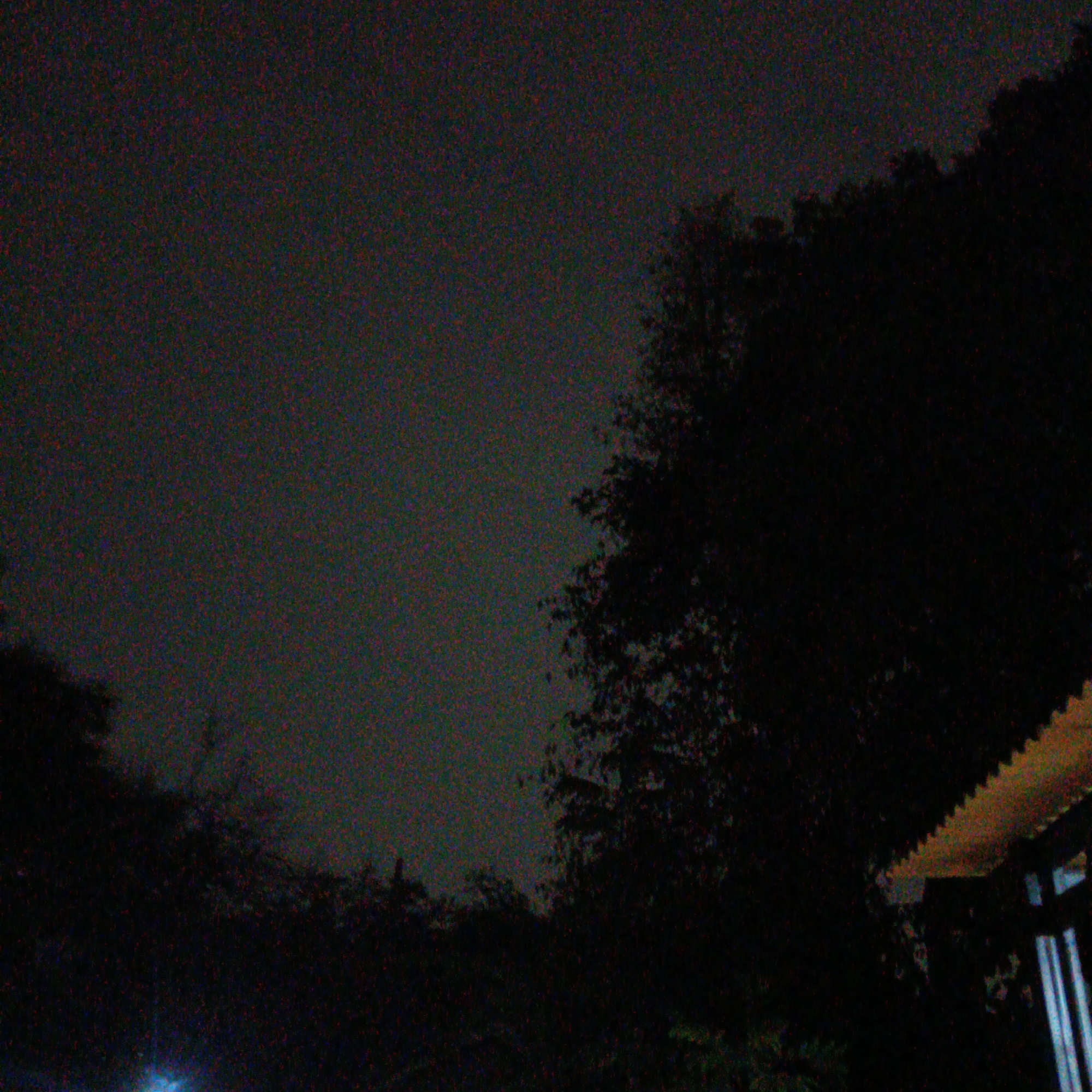The image captures a dark, grainy nighttime scene, likely taken in Jayasampurna, Indonesia. The sky is a muted gray, heavily speckled with digital noise. Silhouetted against the sky are the dense canopies of trees, their forms indistinct in the low light. On the right side of the frame, a section of a building's roof is visible, characterized by a textured, yellowish surface and a corrugated edge. Below the trees, the ground is obscured by shadow. A faint blue light source is detectable in the lower left quadrant, its origin unclear. The overall impression is one of a quiet, nocturnal urban or suburban environment.
