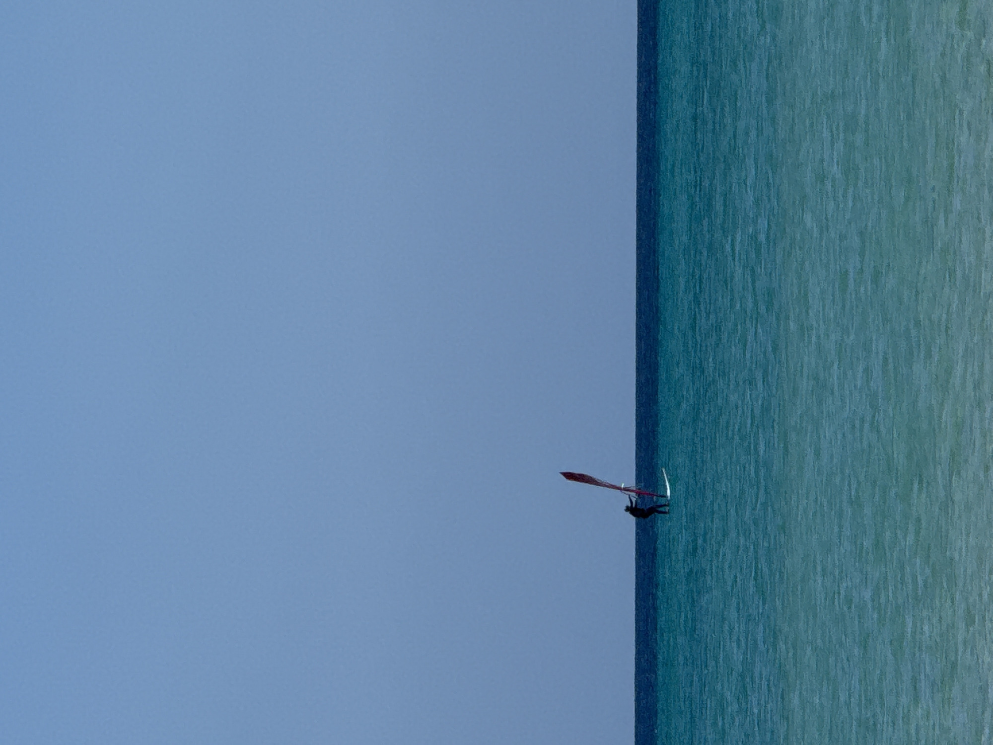 The image displays a lone windsurfer navigating a vast expanse of water under a clear sky. The composition is vertically oriented, with a smooth, light blue sky occupying approximately two-thirds of the frame on the left, and a body of water taking up the remaining right third. A sharp, vertical line acts as the horizon, cleanly separating the sky from the water.
The water, on the right side, is a vibrant teal and turquoise, showing subtle textures of ripples or small waves across its surface, suggesting a gentle breeze. The sky is a uniform, cloudless light blue, indicating bright, sunny weather.
Centrally located along the vertical horizon line is the windsurfer. The individual is largely silhouetted in dark tones, actively engaged with a windsurfing board and sail. The sail is a prominent feature, colored a distinctive red or maroon, and appears to be catching the wind. The person is leaning into the sail, demonstrating movement and control, though specific facial features or expressions are not discernible. The overall impression is one of serene, solitary recreation.
The scene suggests a warm, bright day, ideal for watersports. There are no other objects, boats, or land visible, emphasizing the open nature of the sea and sky. No visible text is present in the image. The location context of Pájara, Spain, aligns with this coastal activity.