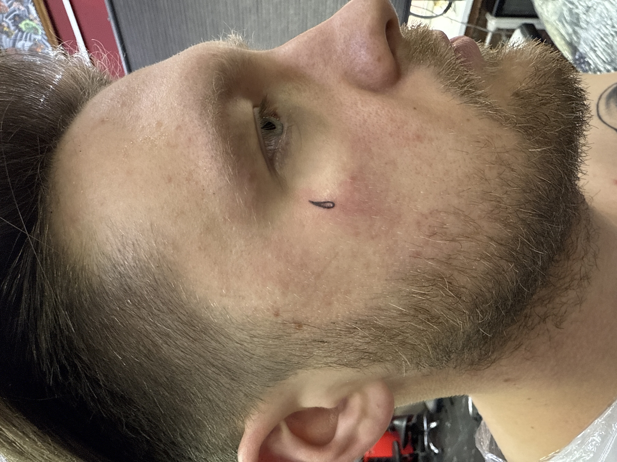 The image presents a close-up, profile view of a young adult male, likely in a tattoo studio in Moscow, Russia. The man is seen from the left side, with his head tilted upwards and to the right, suggesting he is lying down or reclined. He has light skin, light brown or dark blonde hair, and a neatly trimmed, light brown beard. A small, fresh, black, teardrop-shaped tattoo is visible directly under his left eye, surrounded by a subtle redness indicative of recent skin irritation from the tattooing process. His left eye is partially open, and his facial expression appears neutral or relaxed.
Visible around his neck and shoulder is clear plastic wrap, often used to protect clothing or maintain a sterile environment during a tattoo session. The background is blurred and indistinct, showing muted colors and shapes that suggest an indoor setting, consistent with a professional studio environment. There is no visible text in the image. The overall impression is a quiet moment during a tattoo procedure, focusing on the newly applied facial tattoo.
