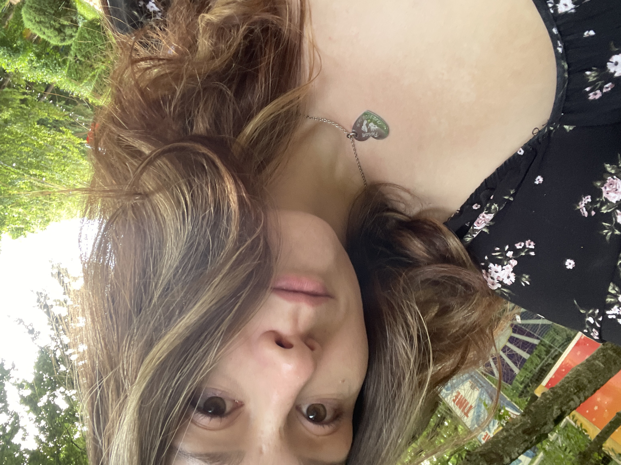A young woman with brown hair and brown eyes is taking a selfie outdoors, with her head tilted to the side. She is wearing a black dress with a floral pattern and a silver necklace with a heart-shaped pendant. In the background, there are trees and what appears to be a sign with the word "TIME" visible. The lighting suggests it is daytime. The woman's facial expression is neutral. The image does not provide specific information about the location within Stavropol, Russia.