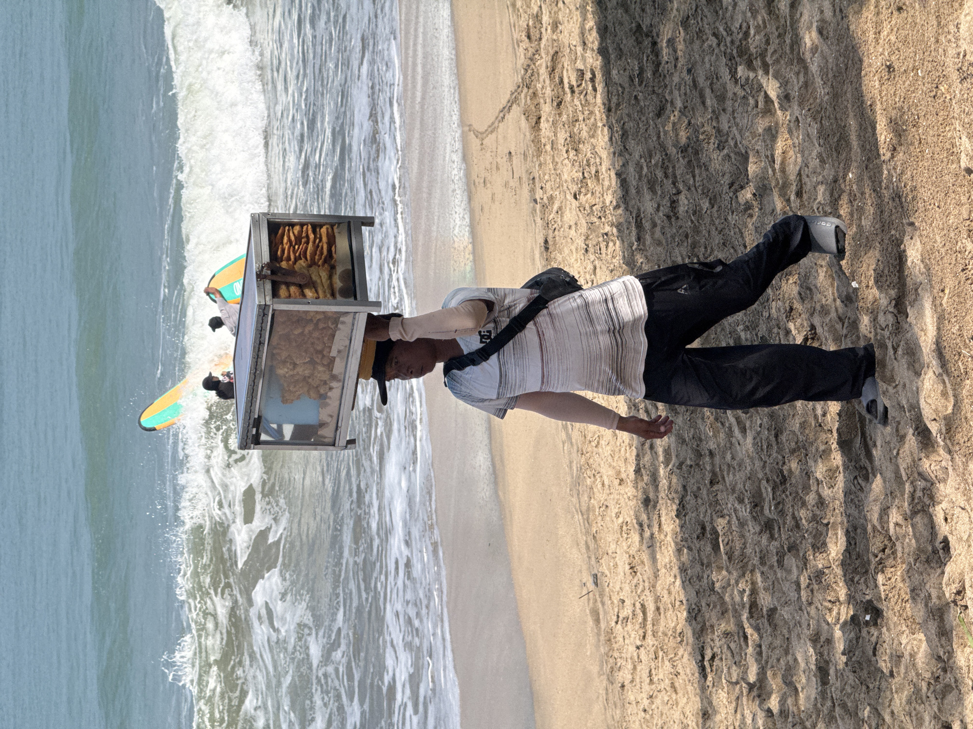A street vendor walks along the beach in Kuta, Indonesia, carrying a food cart on his head. The cart contains various food items, including what appears to be fried snacks and bread. The vendor is wearing a striped t-shirt, dark pants, and sandals. In the background, the ocean waves are crashing onto the sandy shore, and two surfboards are visible in the water. The sun is shining, casting shadows on the beach.