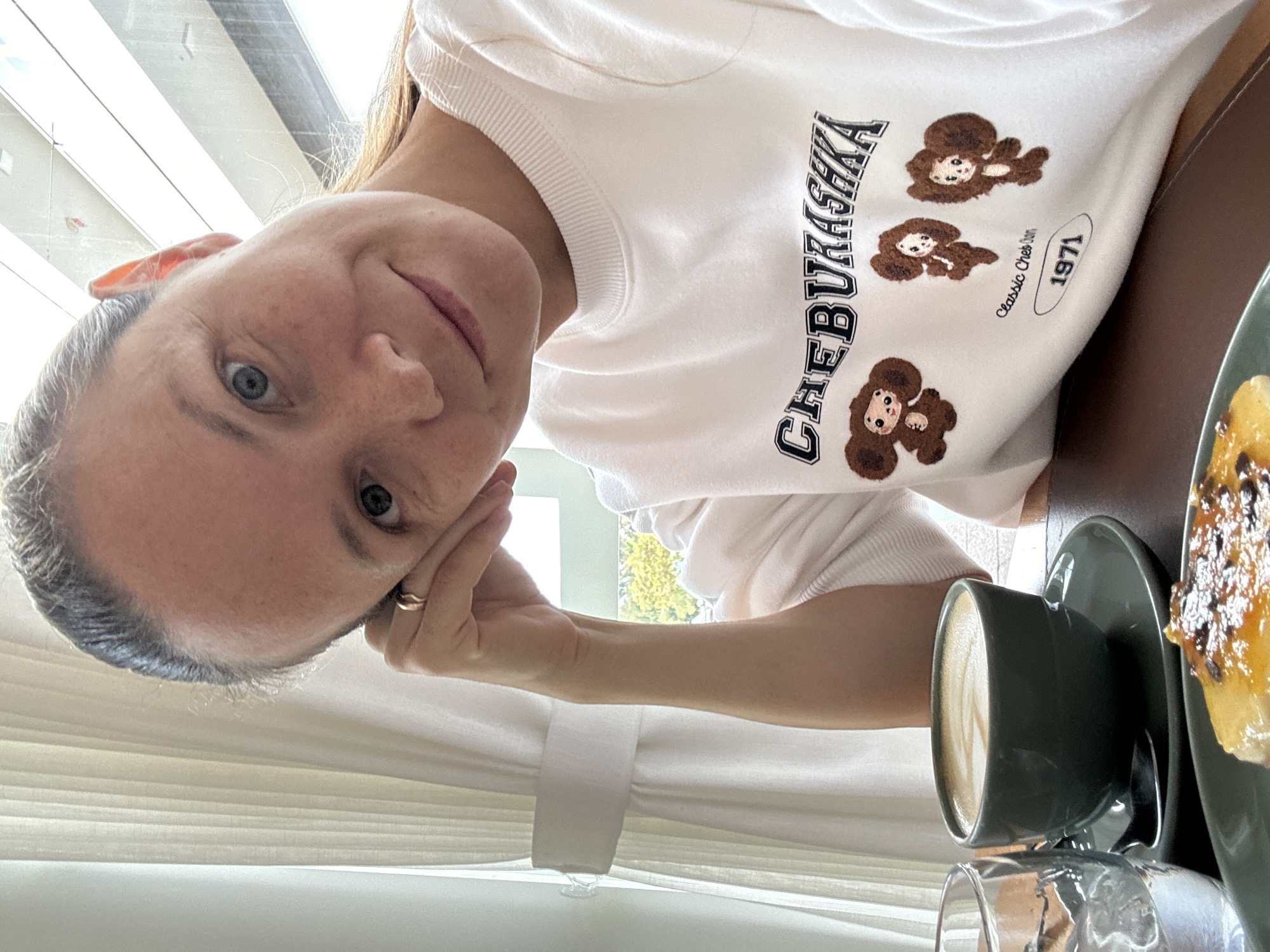 A young woman with light skin, blue eyes, and light brown hair tied back is looking at the camera, resting her head on her hand. She is wearing a white sweatshirt with "CHEBURASHKA" and images of three cartoon characters printed on it, along with "Classic Cheb" and "1971". In front of her is a dark green mug of coffee with latte art, a dark green saucer with a spoon, and a glass of water with ice. A plate with a pastry topped with syrup and chocolate shavings is also visible. The scene appears to be indoors, possibly in a cafe, with light-colored curtains and a window in the background showing trees and foliage. There is no specific information in the image to indicate the location is Florianópolis, Brazil.