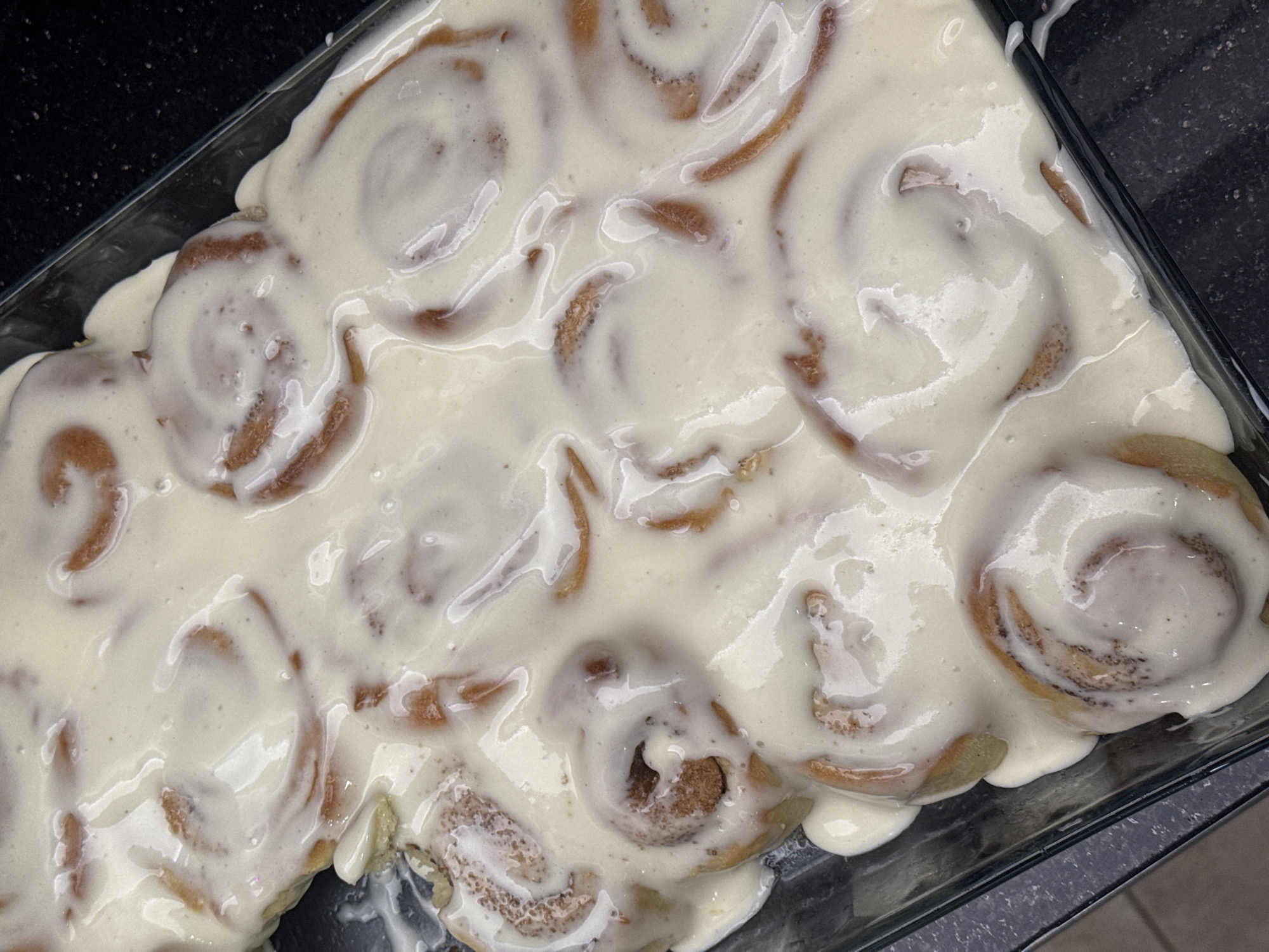 The image shows a rectangular glass baking dish filled with cinnamon rolls covered in a thick, white icing. The rolls are packed tightly together, and the icing is generously applied, covering most of the visible surface. The background is a dark, possibly black, surface, suggesting a kitchen countertop. There is no indication of the location being Yerevan, Armenia.