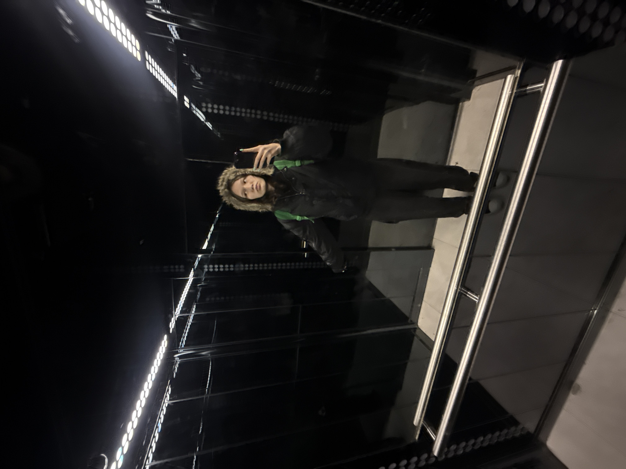 A woman is taking a selfie in what appears to be an elevator. She is wearing a black winter coat with a fur-trimmed hood and a green backpack. The elevator has black, reflective walls and a bright row of lights along the ceiling. There are stainless steel handrails. The location appears to be Saint Petersburg, Russia.