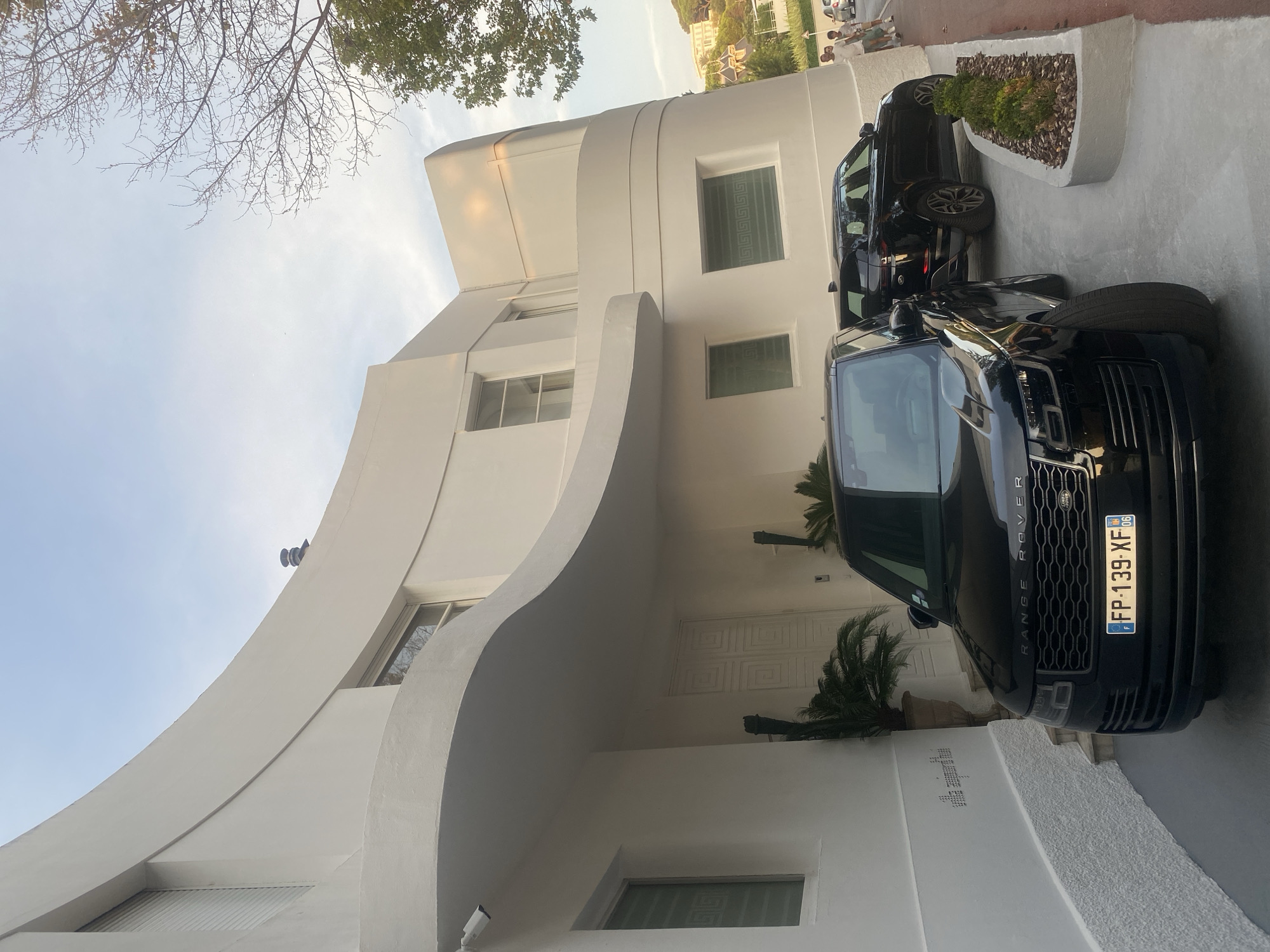 In Antibes, France, two black Range Rover vehicles are parked on a paved driveway in front of a large, white, modern villa. The villa has a distinctive curved architectural design, with smooth, white exterior walls and large windows. The front Range Rover is a newer model, facing forward with its license plate "FP-139-XF" clearly visible. Behind it, a second black Range Rover is parked, angled slightly away from the viewer. The scene is illuminated by natural daylight, casting soft shadows. The driveway is bordered by a planter box filled with green shrubbery. In the background, a few people are walking on a sidewalk. Trees and other buildings are visible in the distance, suggesting an urban or suburban setting. The sky above is a pale blue with some wispy clouds.