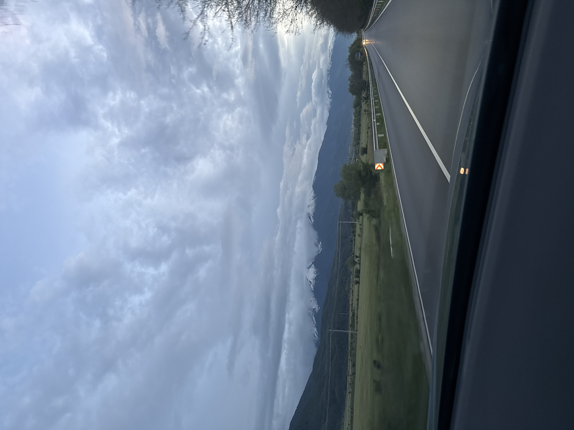 A view from inside a vehicle, presumably a car, looking out onto a highway with mountain views. The sky is overcast with dense clouds. The mountains in the distance are dark with patches of snow. A green field borders the highway, and utility poles are visible. On the highway, a white line divides the lanes, and a yellow arrow sign is seen on the side. Another vehicle is driving in the distance. The location of the scene is Unknown, Unknown.
