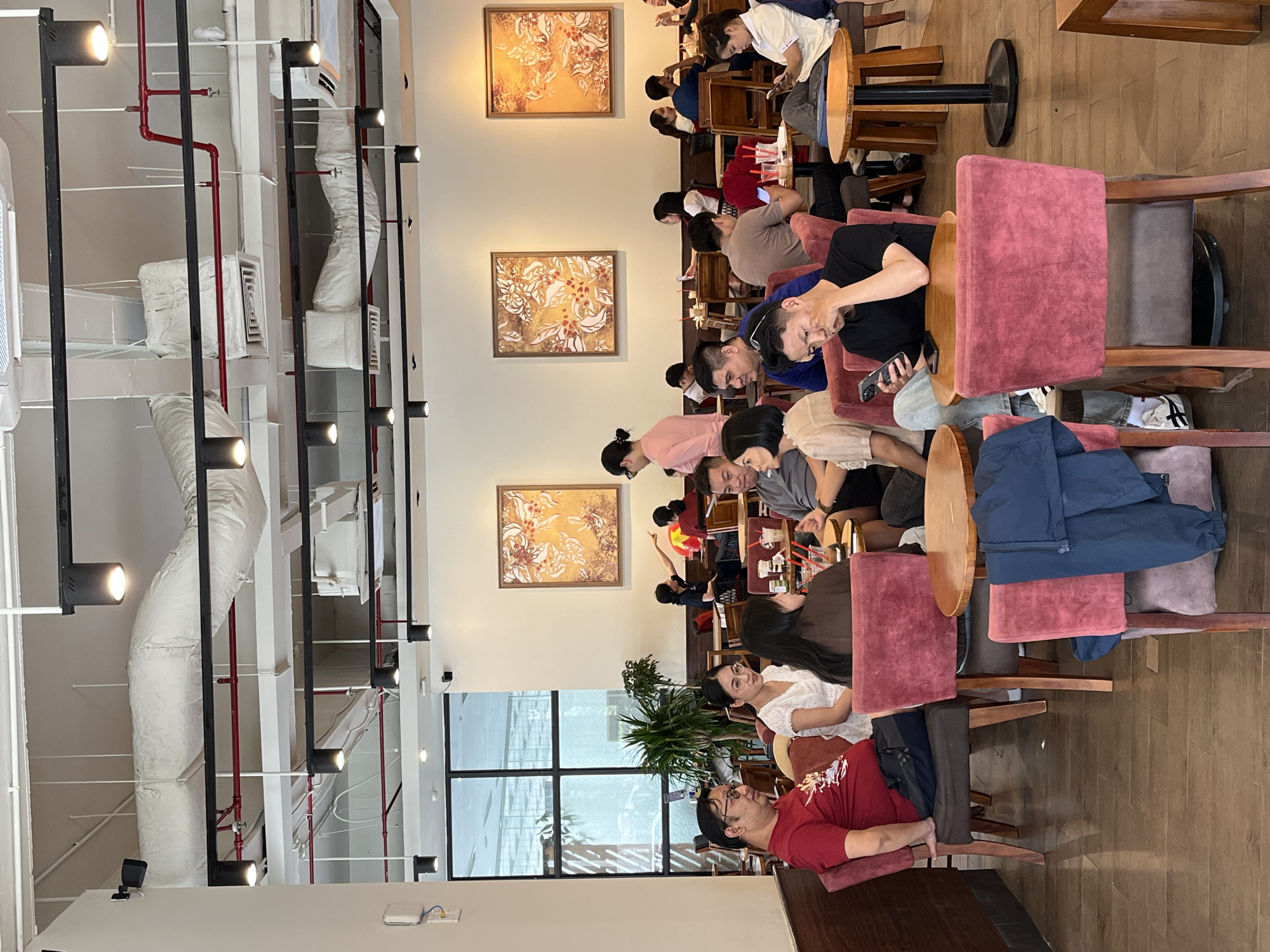 This image captures a vibrant indoor cafe or casual restaurant scene, consistent with a setting in Đà Nẵng, Vietnam. The space is filled with numerous people, primarily young adults, gathered in groups around various wooden tables.
The subjects are mostly of East Asian descent, engaged in a mix of social interaction and personal activities. Many individuals are seen looking at their mobile phones, while others appear to be conversing. In the foreground, a man in a red shirt and glasses is seated, resting his arm on a wooden table. Nearby, a woman in a white top and glasses looks forward or towards another person. Further back, a man in a blue shirt with glasses is also looking down, possibly at a device. Drinks are visible on some tables, suggesting patrons are enjoying beverages or light refreshments.
The setting boasts a modern interior design with an industrial aesthetic on the ceiling, featuring exposed white air ducts, red pipes (likely part of a fire suppression system), and black track lighting fixtures with multiple spotlights. The walls are light-colored, adorned with two framed artworks depicting intricate gold, orange, and white floral or leafy patterns. Large windows on one side offer a glimpse of an urban exterior, implying it is daytime. The floor is covered with warm wooden planks, and the seating includes wooden chairs with both plain and upholstered cushions, some in a distinctive pink or red hue.
The overall atmosphere suggests a relaxed, social gathering. No discernible text is visible anywhere within the frame.