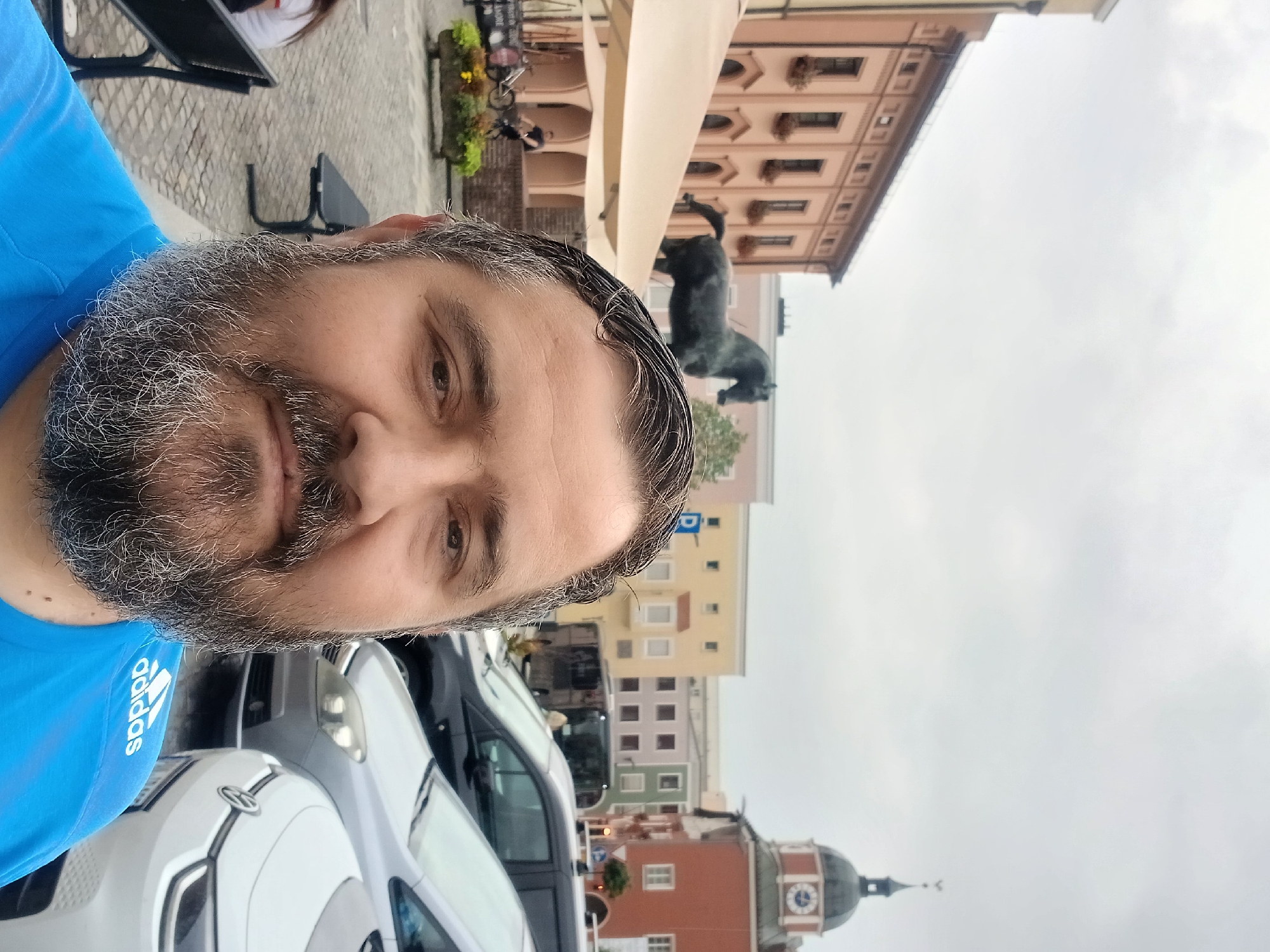 The media file is a vertical, low-angle selfie of a man with a beard, wearing a blue t-shirt with the "adidas" logo visible on the sleeve. The man is in the foreground, with his face filling a significant portion of the frame. Behind him, the scene depicts an outdoor setting with several cars parked, including a white electric car. In the background, there are buildings with colorful facades, suggesting an urban or town environment. One building has a prominent clock tower. A statue of a black animal, possibly a dog or a panther, is visible on a pedestal in front of one of the buildings. The sky is overcast, indicating a cloudy day. The overall impression is a casual snapshot taken during the daytime, likely in a European town square or street given the architecture and parked vehicles. The man's expression is neutral. Visible text includes the "adidas" logo on his shirt and a license plate on one of the cars. The presence of the clock tower and the style of the buildings suggest the location could be in Germany.