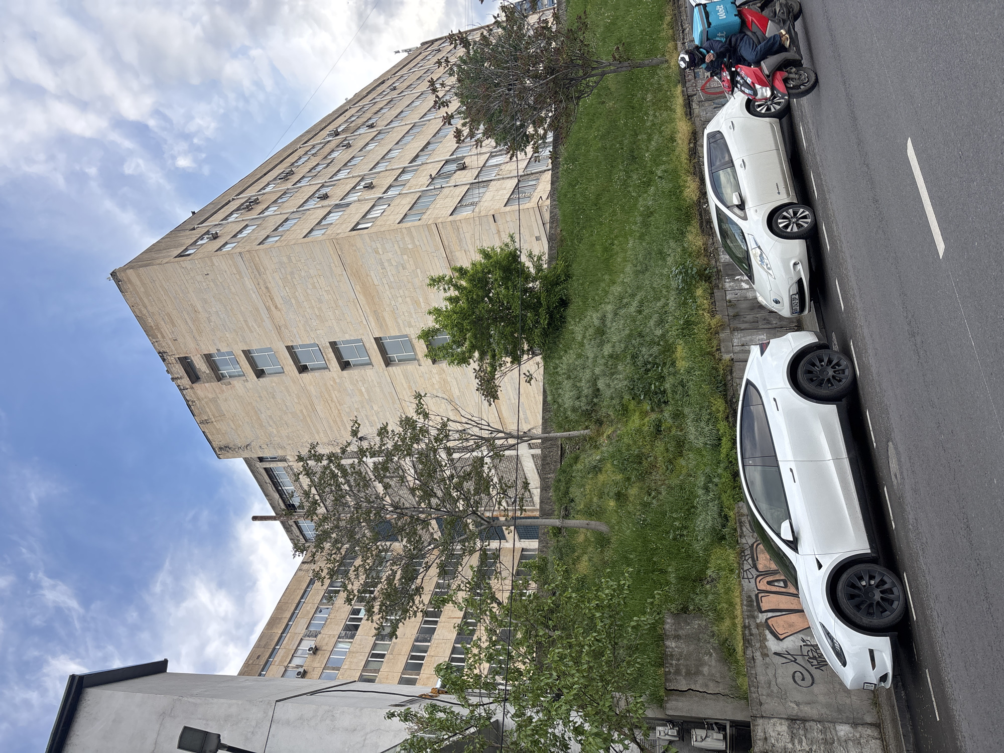 The image shows an urban street scene in Tbilisi, Georgia. A tall, aged building with a beige facade dominates the left side of the frame, its multiple windows suggesting it is an office or apartment building. In front of the building, a strip of grassy area with small trees separates the building from the street. Parked along the right side of the street are two white cars, and a red scooter with a rider wearing dark clothing and a helmet is seen behind the first car. The sky is partly cloudy. Graffiti is visible on a low wall near the bottom of the image. The scene seems to be taken during the daytime.