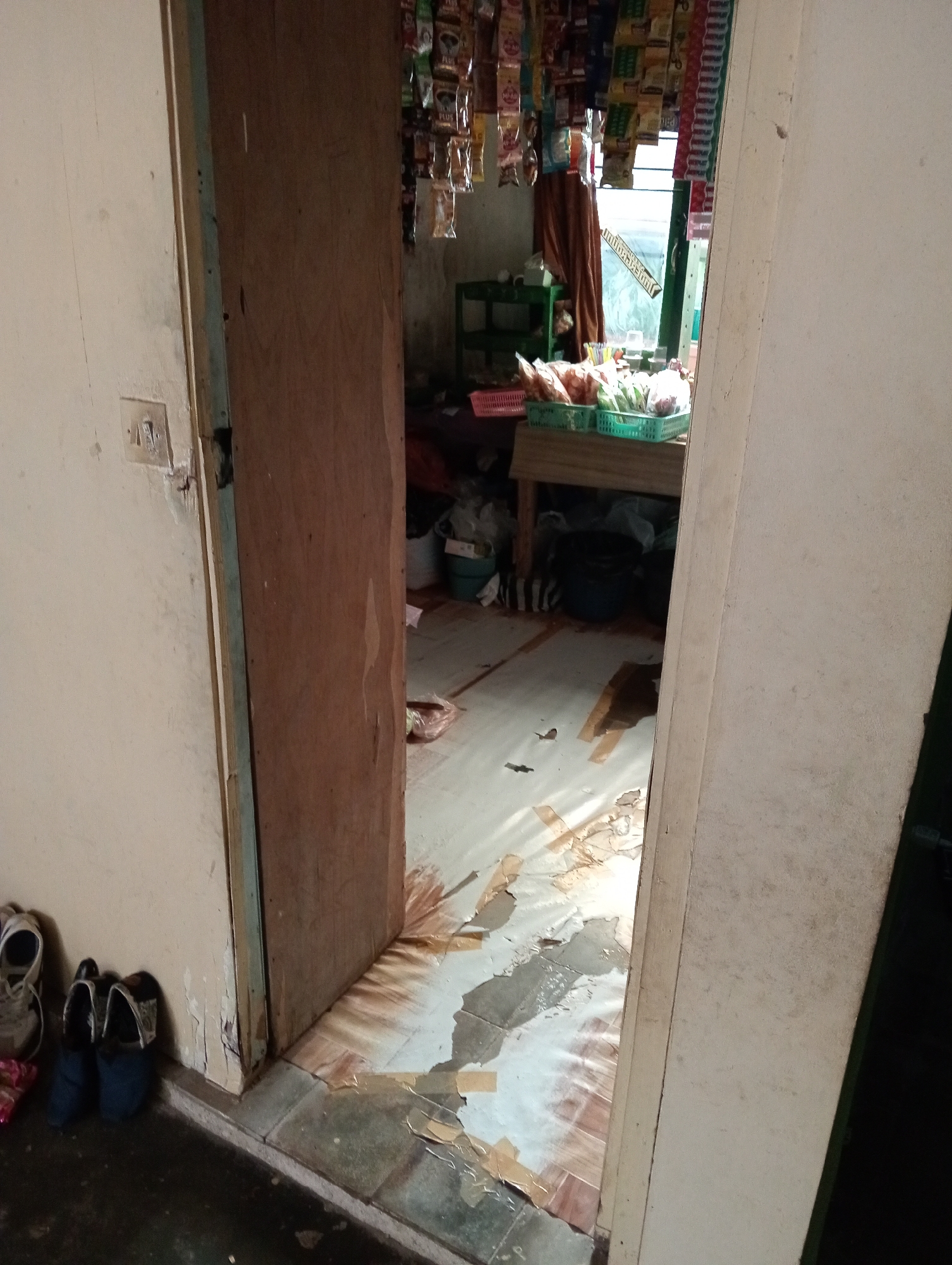 The image shows an interior view of a room that appears to be a small shop or convenience store, likely located in Krajan Kidul, Indonesia. The foreground features a doorway with a wooden door slightly ajar, revealing a glimpse into the interior. To the left of the doorway, near the wall, are several pairs of shoes, including a pair of white athletic shoes and two pairs of blue sandals. The wall on the left has a light-colored paint and a visible light switch.
The interior of the room is cluttered. Shelves are visible, stocked with various packaged goods hanging from above. A green shelf unit holds some items, and in front of it, green plastic baskets are filled with what appear to be snacks or other merchandise. A window is present in the background, covered partially by a brown curtain. The floor inside the room has some debris and peeling linoleum or floor covering. There are buckets and bags visible on the floor as well, adding to the general disarray. The overall impression is of a busy, lived-in space, characteristic of a small retail establishment in a residential area.