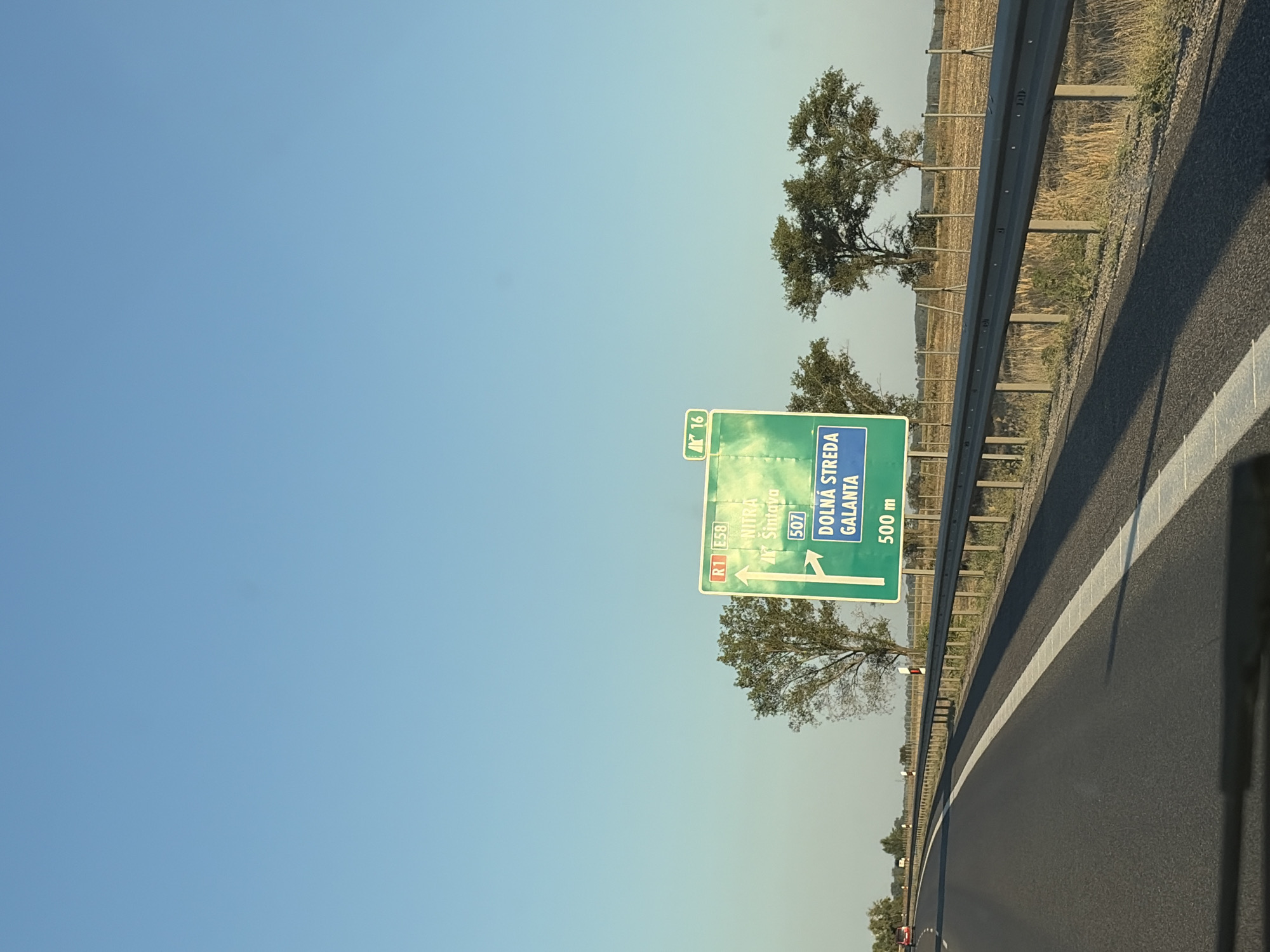 An outdoor, eye-level, wide shot shows a highway sign in Dolná Streda, Slovakia, on a clear, sunny day. The sign is green with white and blue text and arrows indicating directions. Text on the sign reads "R1 E58" and "16" in smaller boxes. Below this, it lists "NITRA" and "Šintava" with an upward arrow, and "507" in a box. Further down, it says "DOLNÁ STREDA" and "GALANTA" with a right-pointing arrow, and "500 m" below that. The highway itself is visible in the foreground, with a white line on the left side. A metal guardrail runs along the side of the road, separating it from a dry, grassy field. Several trees are scattered throughout the landscape, with a few more visible in the background. The sky is clear and blue.
