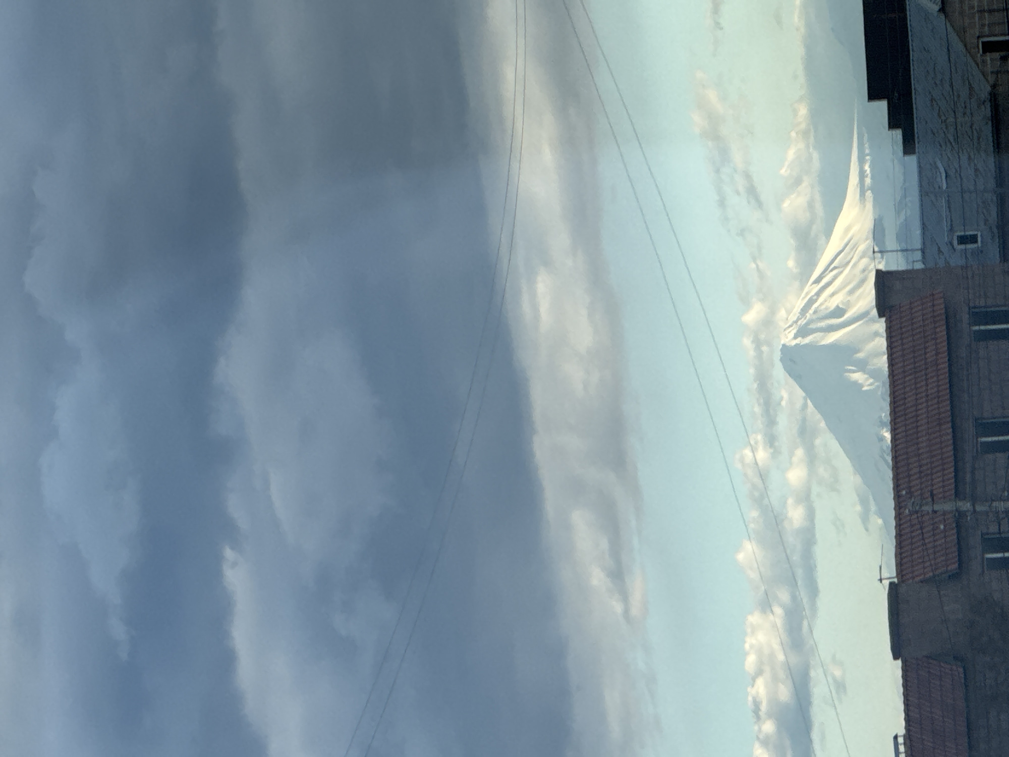 An eye-level, vertical shot, taken in Verin Ptghni, Armenia, shows a cloudy sky to the left with power lines running across it, while a mountain can be seen on the right. Building structures are visible in the lower right corner of the frame. The mountain's peak is snow-capped and contrasts against a sky with scattered clouds.