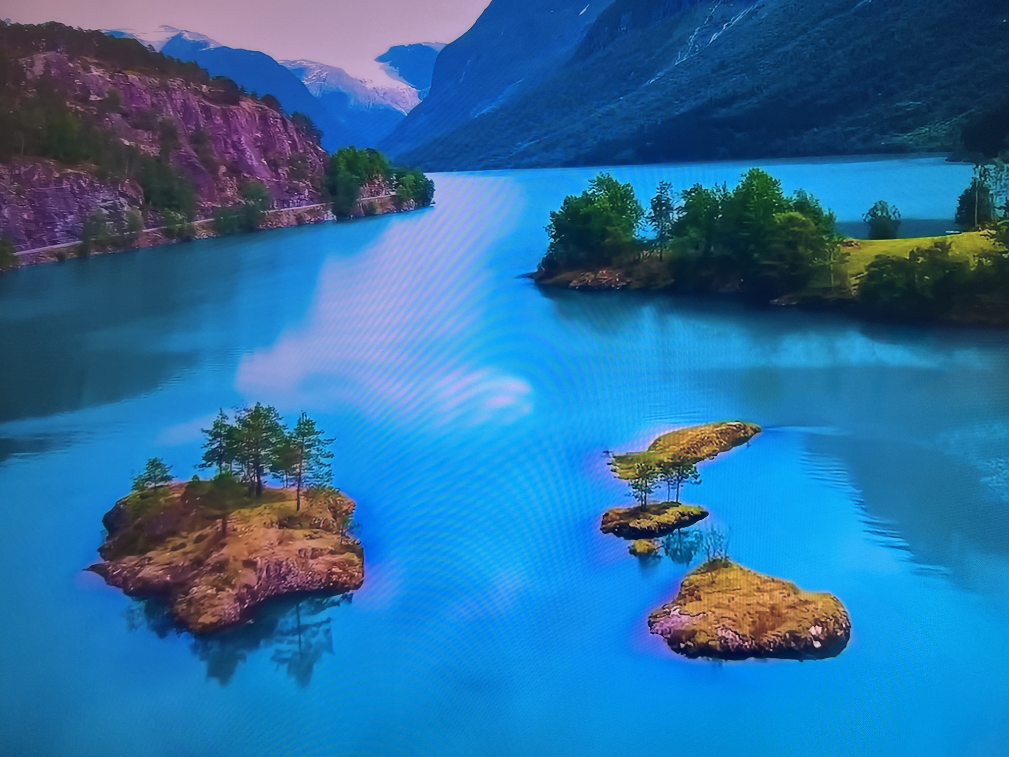The image depicts a serene landscape of a large body of water surrounded by mountains and islands. The water is a vivid blue. On the left side of the frame, a mountain with exposed rock and green vegetation rises, with a road visible along its base. In the background, snow-capped mountains can be seen. The right side of the frame shows darker mountains descending to the water's edge. Several small, tree-covered islands dot the water. The islands vary in size and shape, some with a single tree or a small cluster of trees.
This image is most likely from somewhere in Norway, as this is a very typical landscape from the Nordic country.
There is no evidence that this image is related to Basingstoke and Deane, United Kingdom.