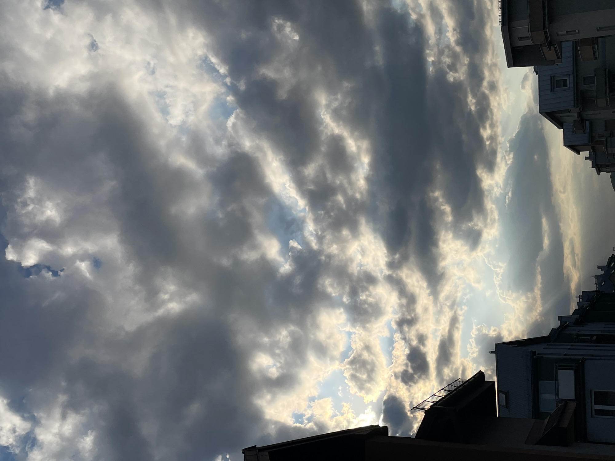 The image depicts a sky filled with dark, striated clouds with sun rays peeking through. The clouds are streaked across the sky, with variations in grey and white tones. Below the clouds, rooftops and buildings are visible, suggesting an urban environment. The buildings' architectural style appears modern, with angular rooftops and visible windows. The image has a slightly upward angle, emphasizing the height of the clouds and buildings.