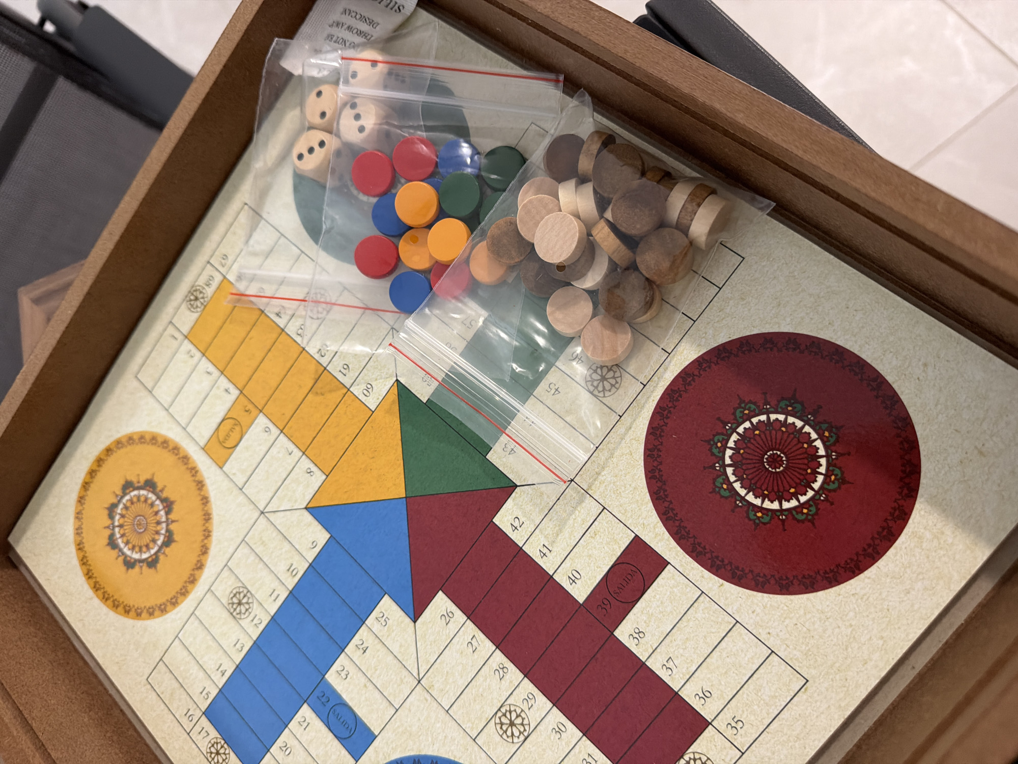 A high-angle, slightly blurry, close-up shot shows a wooden board game, likely Ludo or a similar game, in a brown wooden box. The game board features a cream-colored surface with a grid of numbered squares and colored pathways, including blue, orange, green, and red. Decorative circular motifs are present on the board. Several plastic bags containing game pieces are scattered on the board. One bag holds six dice, each showing various pips. Another bag contains an assortment of circular game tokens in red, blue, and orange colors. A third bag holds numerous wooden tokens, some appearing to be stacked. The game board is positioned within the confines of the brown wooden box, which is set against a neutral background. The scene is indoors, but the specific location in Finestrat, Spain cannot be determined from the visual information provided.