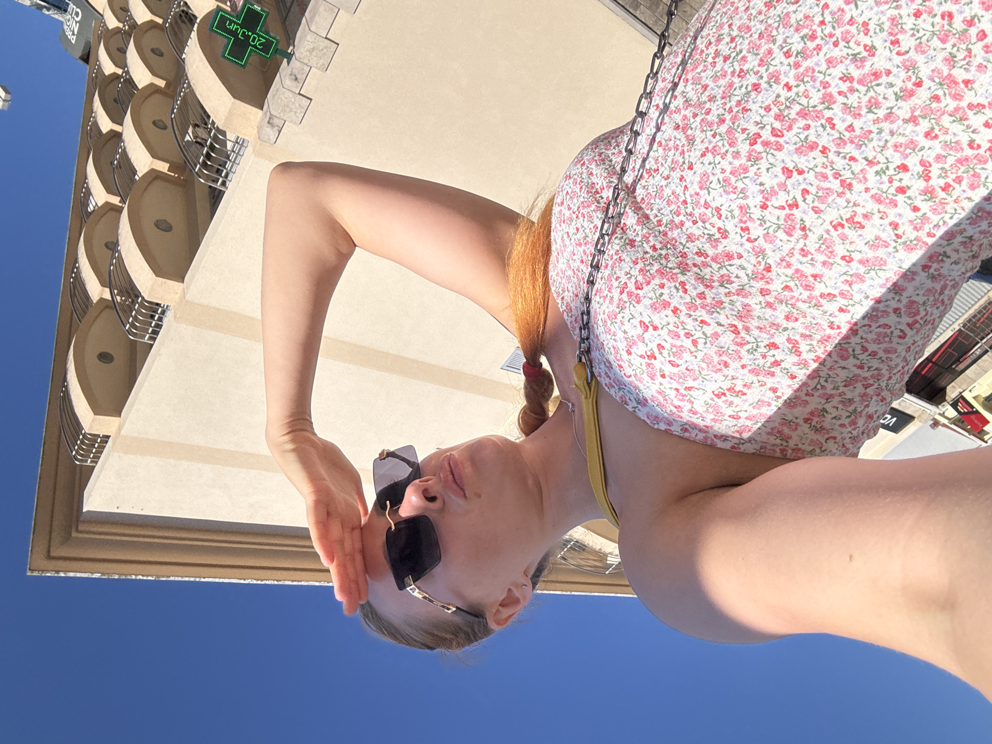 A low-angle, full shot captures a blonde woman from the chest up, outside on a sunny day, in front of a tan-colored building. The woman is wearing oversized, dark sunglasses and a floral print dress with a yellow strap visible over her shoulder. Her right arm is raised, with her hand shielding her eyes from the sun as she looks upward. Her blonde hair is braided into a pigtail. Behind her, the building features a repeating pattern of arched balconies with metal railings. A green cross-shaped sign with the text "20.3°C" is visible on the right side of the building. The sky is a clear, bright blue. The location is identified as being in the city of Budva, Montenegro.