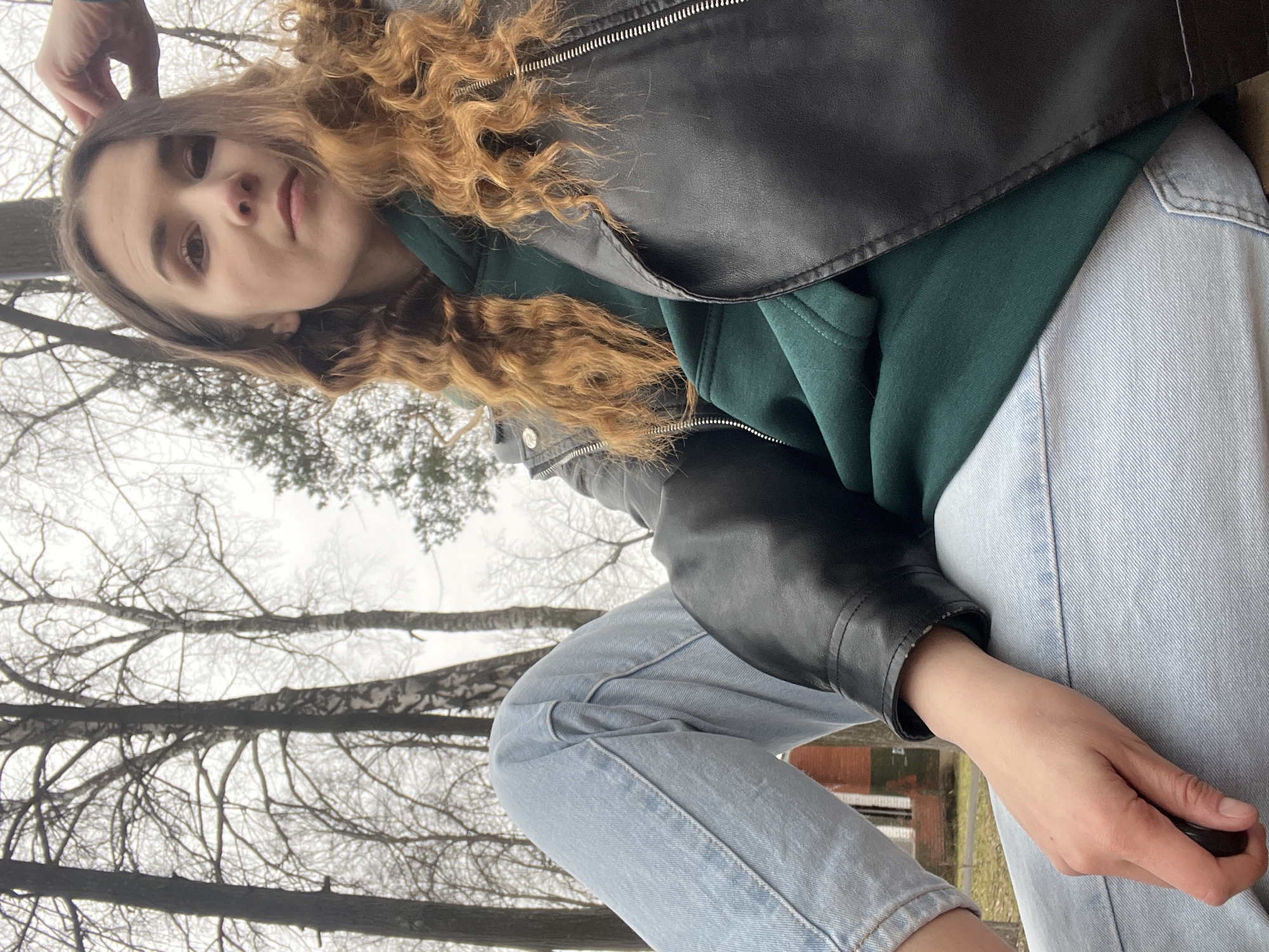 The image shows a person in an outdoor setting with bare trees visible in the background, suggesting a cold season like late autumn or early spring. The person has curly, reddish-brown hair and is wearing a dark green hoodie under a black leather jacket and light blue jeans. The composition is from a slightly upward angle. The background shows birch trees. While it is not possible to determine the specific location within Votkinsk, Russia, the bare trees and architectural elements vaguely visible in the background might indicate a park or a less densely populated area of the city. The person is holding their hand to their head.