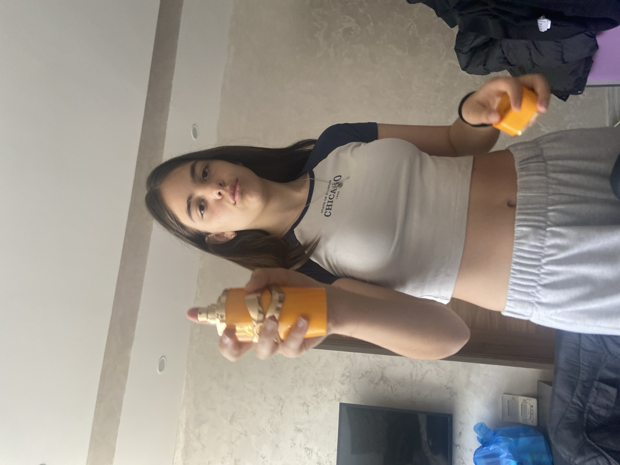 A teenage girl is indoors, holding a yellow perfume bottle in her left hand and a yellow object in her right. She is wearing a white and navy blue top with the word "CHICAGO" on it and grey sweatpants. The background includes a patterned wall, a TV, and possibly clothing. There is no confirmed evidence to associate this picture with the city of Dragovishtitsa, Bulgaria, and therefore I can not provide location details.