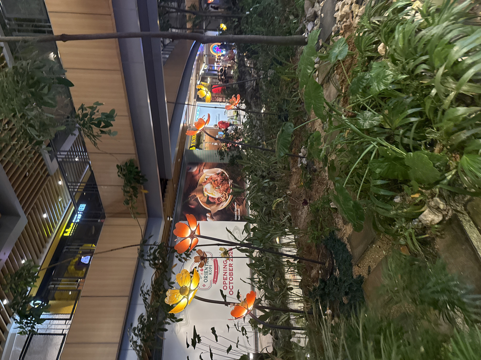 The image depicts an indoor botanical display, likely within a shopping mall in Kuala Lumpur, Malaysia, given the presence of signage for "ORIENTAL KOPI," a local Malaysian coffee chain. The foreground features a lush, tropical-themed garden with various plants and artificial flowers that appear to be illuminated from within, emitting a warm orange and yellow glow. These illuminated flower sculptures are the most prominent elements in the foreground. In the midground, a large banner advertises the opening of "ORIENTAL KOPI" in "OCTOBER 2023." Behind this, a balcony level of the mall is visible, with shops and people. The architecture suggests a modern shopping complex with multiple levels and wooden paneling. The overall atmosphere is one of artificial greenery and commercial branding, common in urban shopping environments.