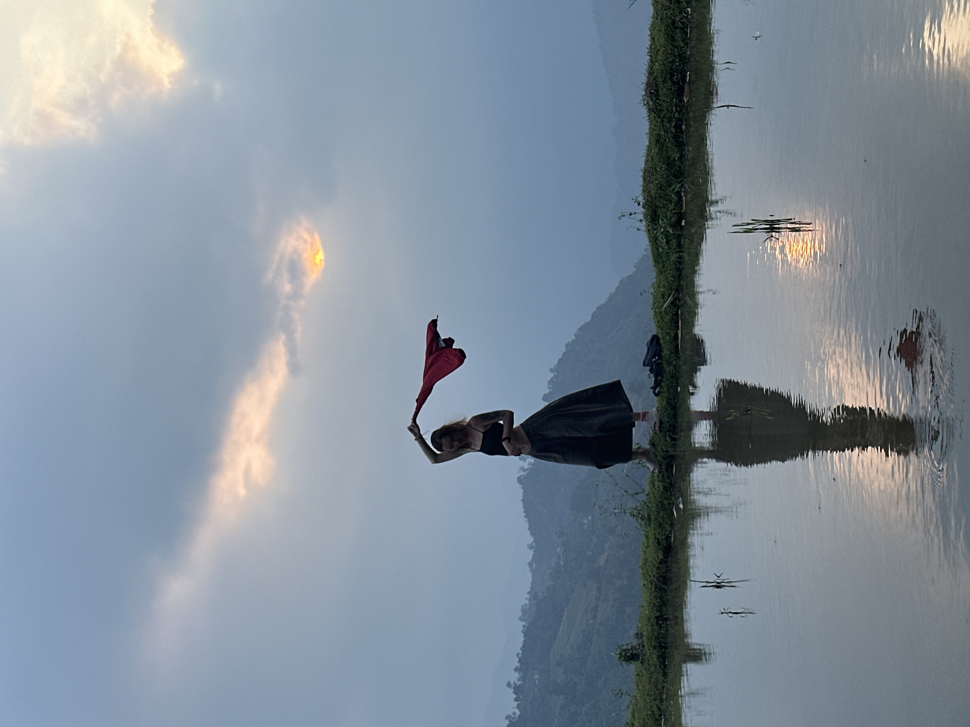 The image captures a serene outdoor scene, likely at sunset or sunrise, featuring a woman standing beside a body of water. The woman, positioned slightly left of center, is dressed in a dark top and a long, dark skirt. She stands on a narrow strip of green land or grass that separates her from the water. Her arms are raised, and she holds a vibrant red piece of fabric that billows upwards and to her left, suggesting it's caught by a gentle breeze.
The still water to her right acts as a mirror, clearly reflecting the sky, clouds, and the prominent golden-orange light of the sun. The actual sun is partially visible through a break in the clouds in the upper left portion of the sky, casting a warm glow. Its reflection on the water creates shimmering streaks of light that extend towards the middle and lower right of the frame.
In the background, a series of hazy, dark green mountains rise, their forms softened by the ambient light and atmosphere. The sky above is a pale blue, with scattered clouds that range from soft white to a light grey. The overall setting is natural and tranquil, characteristic of a rural landscape, such as a flooded rice paddy or a calm lake. There is no visible text in the image. This scene evokes a peaceful moment in a beautiful natural environment, consistent with the given context of Nậm Dịch Commune, Vietnam.