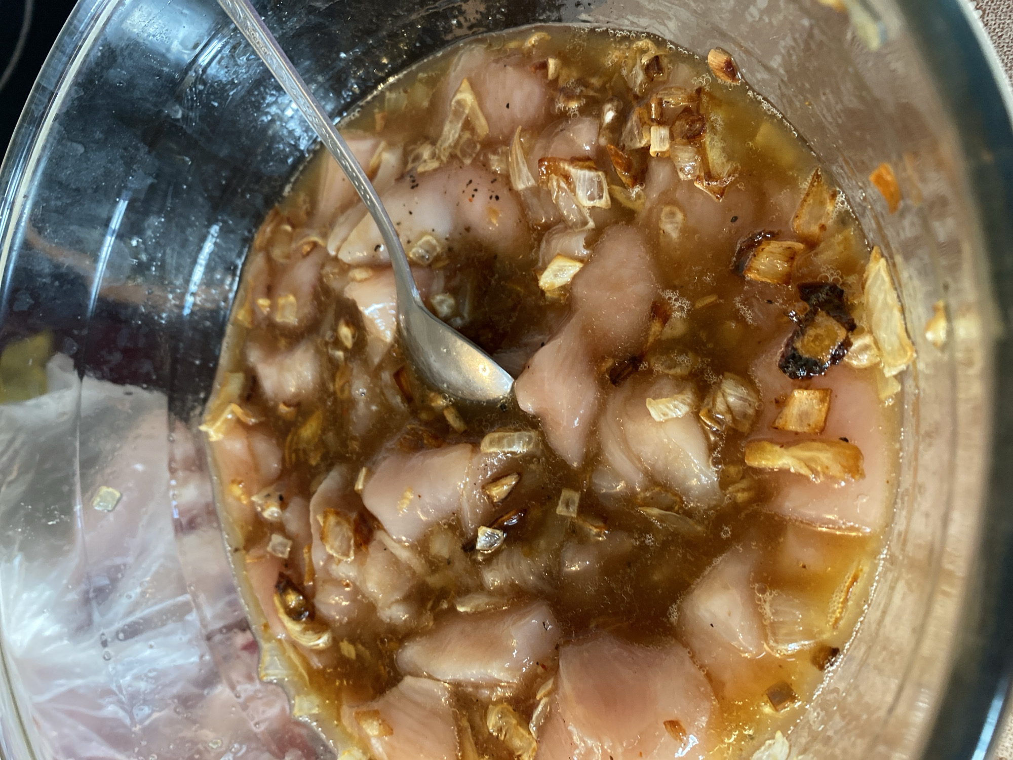 A close-up, overhead shot shows a glass bowl containing raw, cubed chicken in a marinade. The marinade is a light brown liquid with chopped onions and spices, including black pepper. A silver spoon rests in the bowl, partially submerged in the marinade. The bowl appears to be on a countertop. There are no people visible in the image. The scene is indoors, with the lighting suggesting it is daytime.