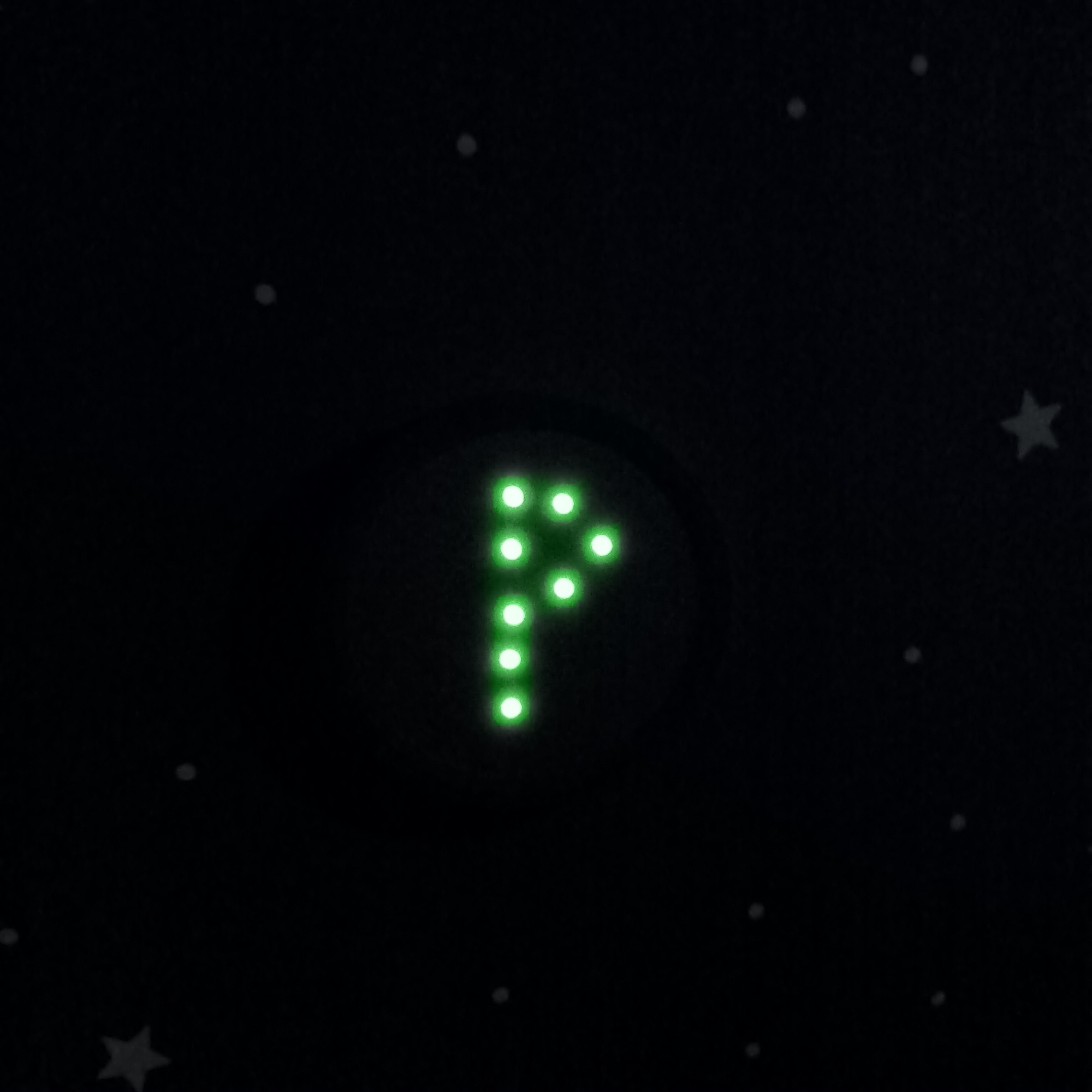 The image shows a dark, star-filled background with a prominent green symbol in the center. The symbol is formed by an arrangement of green LED lights, resembling the letter "P" or a rightward-pointing arrow. Scattered across the dark background are small, white dots that appear to be stars, and a few slightly larger, light gray star shapes are also visible. There are no people, objects, or actions depicted in the scene, and no discernible location within the city of Varna, Bulgaria, can be identified from the visual information provided. The scene appears to be a symbolic representation or a decorative element, possibly related to astronomy or a playful design.