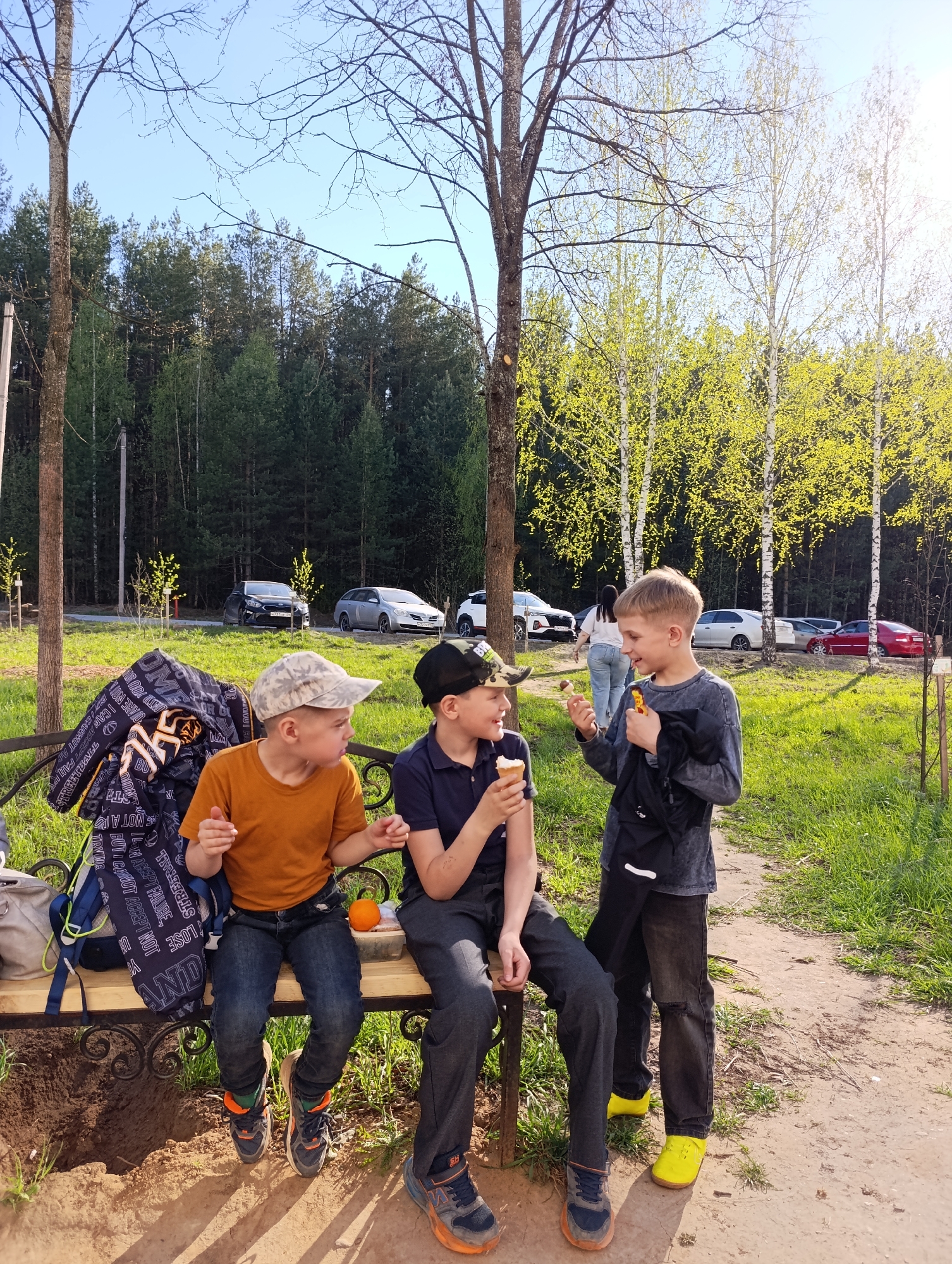 Three boys are outdoors in a park or wooded area in Chaikovsky, Russia. Two are sitting on a wrought-iron bench with a backpack and jacket. The boy on the left wears an orange t-shirt, jeans, and a camouflage cap; he is gesturing with his hands. The boy in the center wears a dark blue collared shirt and dark pants, and he is eating an ice cream cone. The third boy stands on the right, wearing a gray shirt, dark pants, and yellow shoes; he holds an ice cream cone and a black jacket or bag. All three boys are interacting with each other. The background includes a grassy area, trees, and parked cars on a road. The overall setting is sunny and appears to be a park or recreational area in Chaikovsky, Russia.