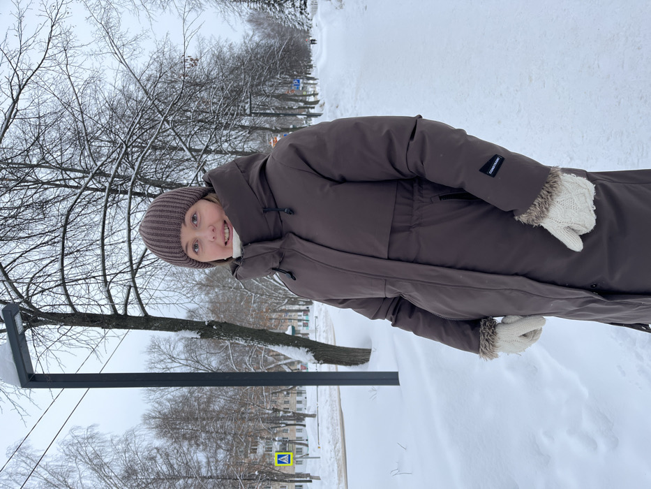 The scene depicts an adult woman standing outdoors in a snow-covered urban setting within Chaikovsky, Russia. She is positioned slightly to the right of the frame's center, facing forward with a faint, closed-mouth smile. The woman wears a dark brown, long, quilted winter coat featuring a high collar, hood, and a cinched waist, with a small black rectangular patch bearing white text, possibly "CONSTANT," on her left sleeve. A light-colored scarf or turtleneck is visible at her neck. She also wears a ribbed dark brown knit beanie and cream-colored cable-knit mittens with faux fur trim.
The environment is uniformly covered in snow. A modern, dark gray street lamp, topped with snow, stands behind and to the left of the woman. In the far left background, a yellow pedestrian crossing sign is visible near a snow-covered street, with multi-story apartment buildings beyond. Bare deciduous trees, also laden with snow, line the background. To the right, a snow-covered pedestrian pathway extends into the distance, with two figures faintly visible further along it. The sky is overcast.