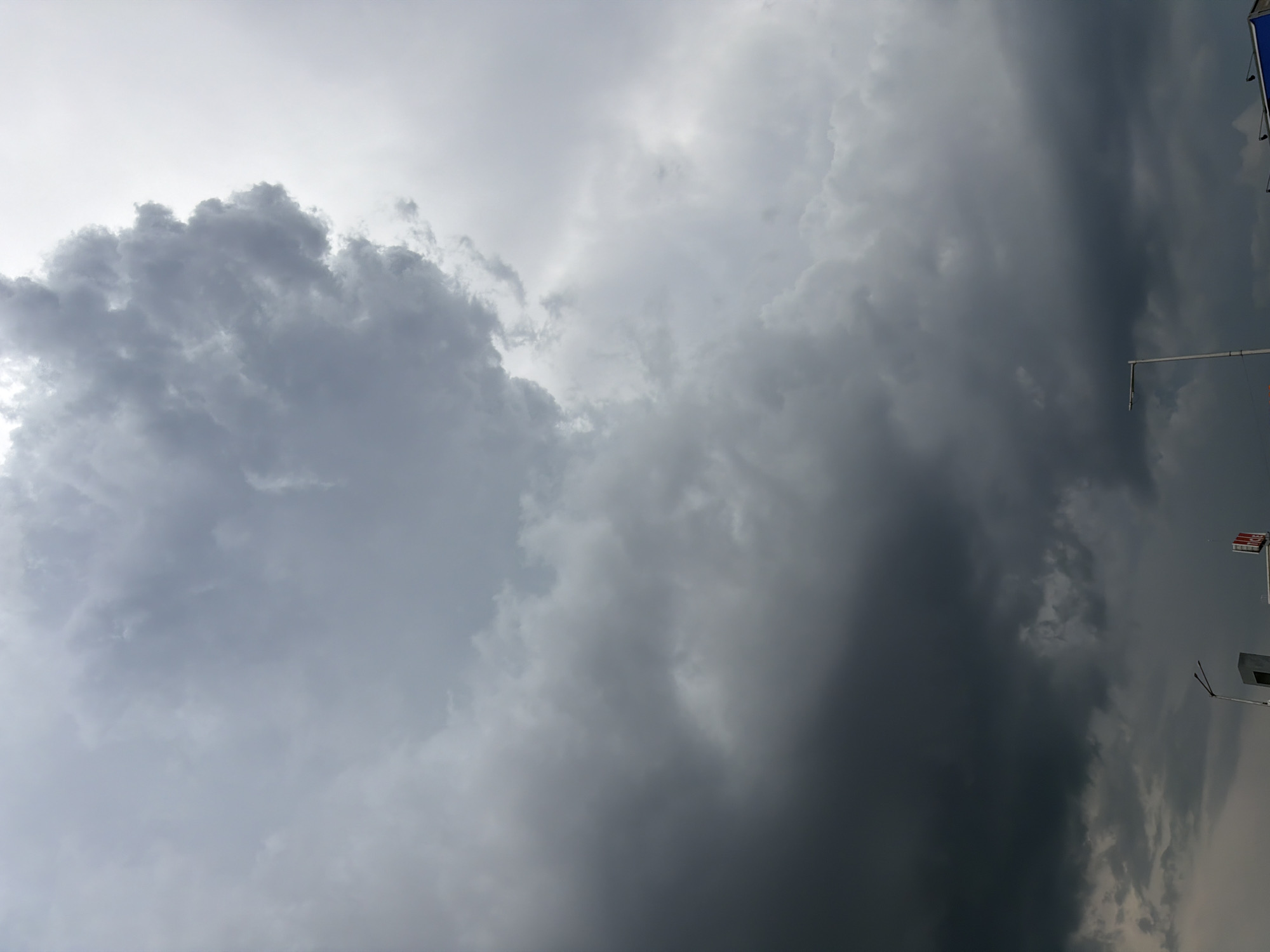 An overcast sky filled with dark, dramatic storm clouds dominates the frame, with lighter clouds and a hint of brighter sky towards the top right. In the lower portion of the image, the tops of some man-made structures are visible, including a streetlight and parts of buildings with signage. The scene appears to be an urban setting, possibly on the outskirts of Sofia, Bulgaria, given the presence of these structures against the backdrop of approaching weather. No people are discernible in the image.