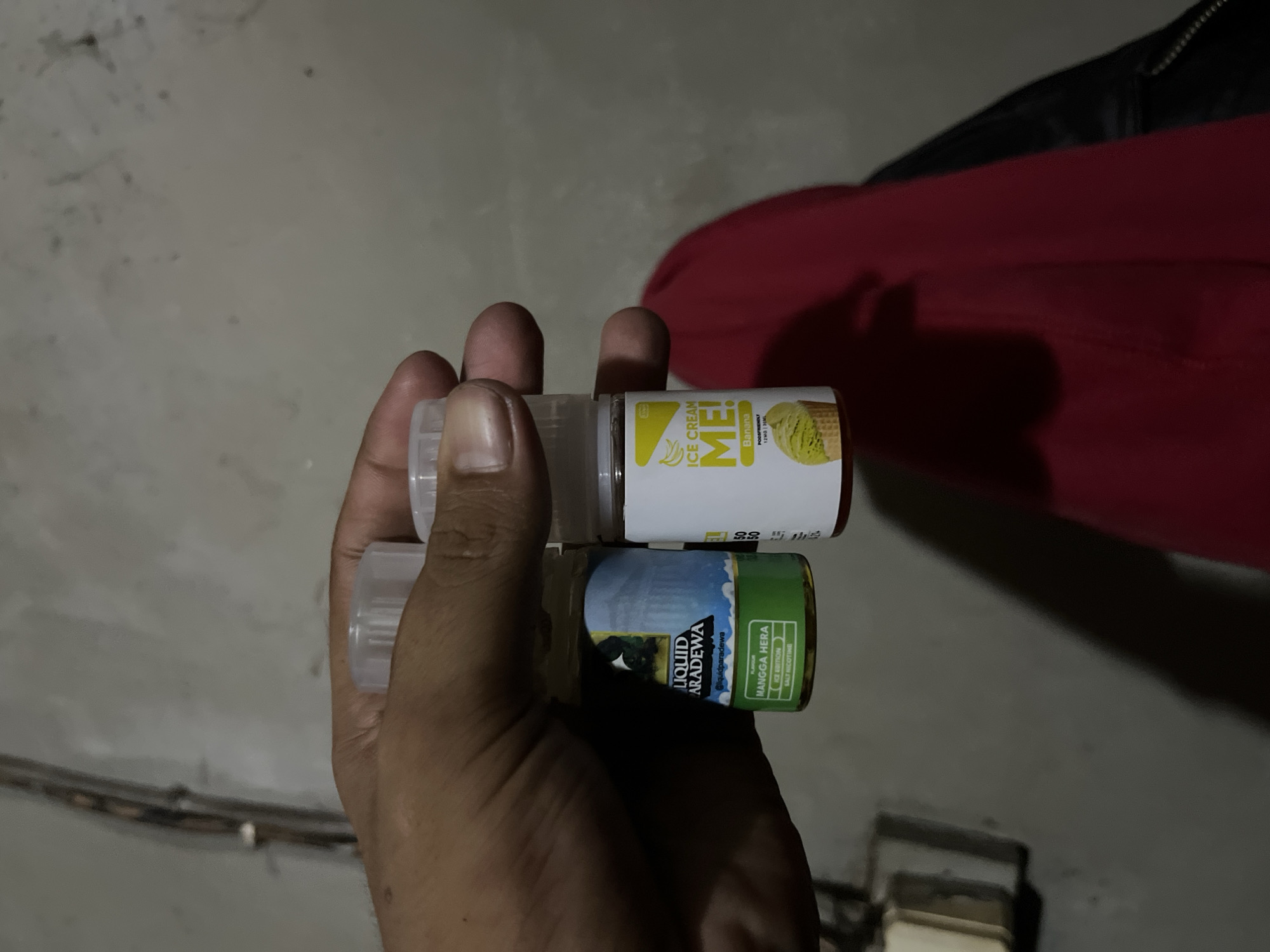 An adult male's hand is holding two small bottles of e-liquid. The bottle on the left is labeled "LIQUID PARADEWA" and appears to have a mango flavor. The bottle on the right is labeled "ICE CREAM ME!" and features a banana flavor. Both bottles have clear plastic caps and are positioned against a blurred background of a wall. The image appears to have been taken indoors. The overall lighting is dim, with the primary light source likely coming from the front, casting shadows on the hand and bottles. There is no indication of the city of Central Jakarta, Indonesia, within the image itself.