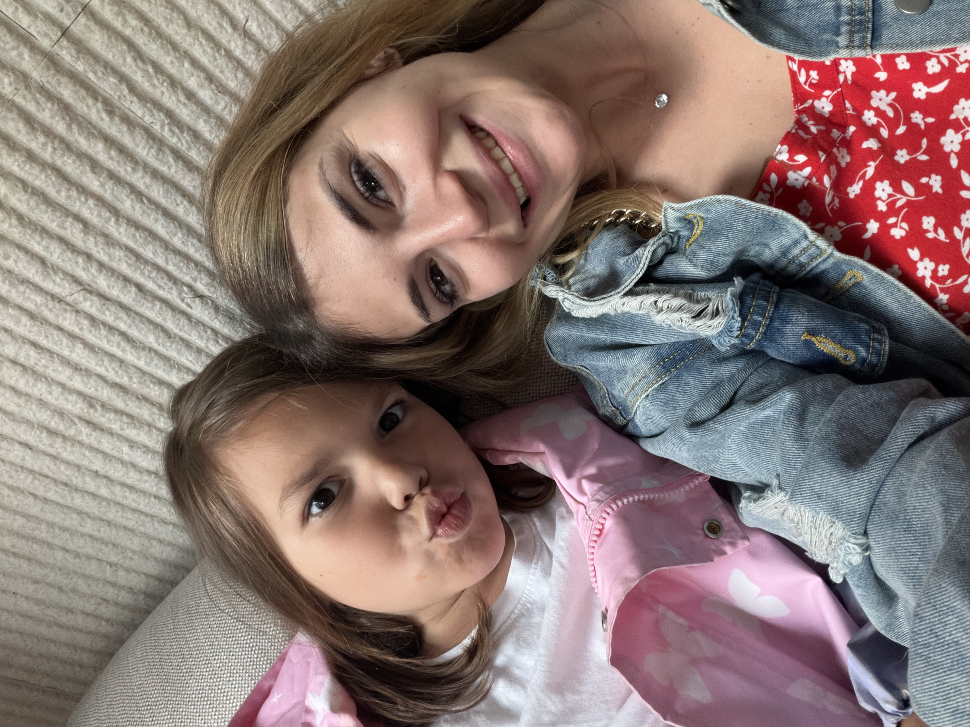 Two people, a woman and a child, are lying down next to each other. The woman is wearing a red floral dress and a denim jacket. The child is wearing a white t-shirt and a pink jacket with white hearts. The background is a beige textured surface. The location is not discernible from the image; there is no visual information suggesting Budva, Montenegro, or any specific city.