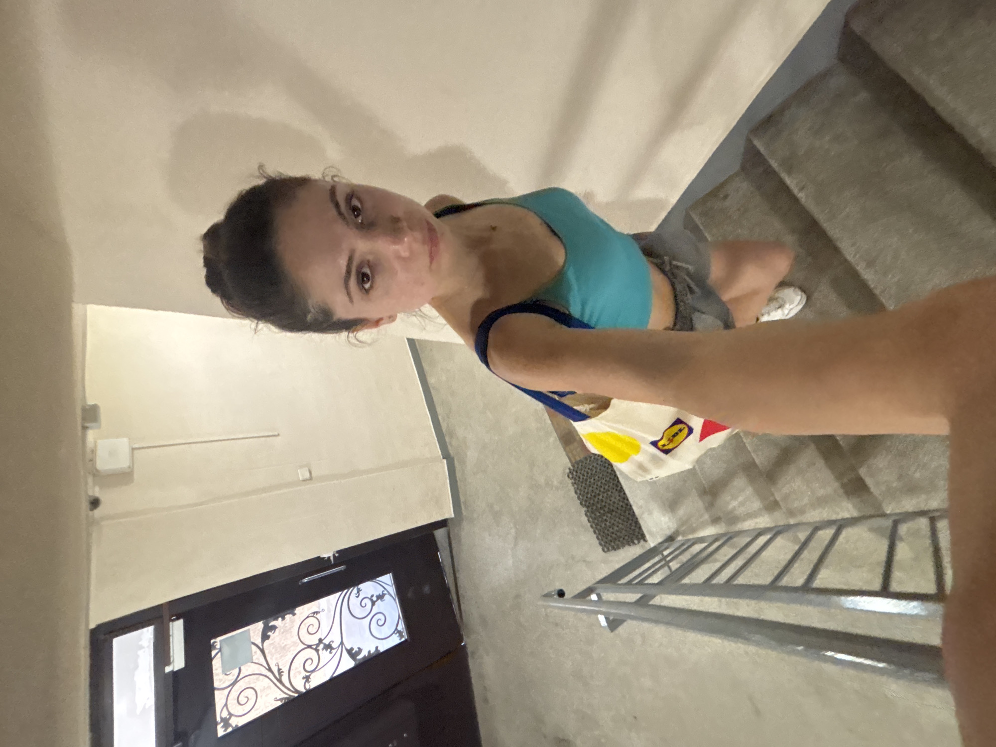 A young woman is taking a selfie on a dimly lit concrete staircase with a metal railing on the left. She is wearing a turquoise tank top and grey shorts, with white sneakers. A white canvas tote bag with yellow and red accents is slung over her right shoulder. The background shows a dark brown door with decorative metalwork and a light-colored wall. The scene appears to be an indoor stairwell in a residential building, possibly in Varna, Bulgaria.
